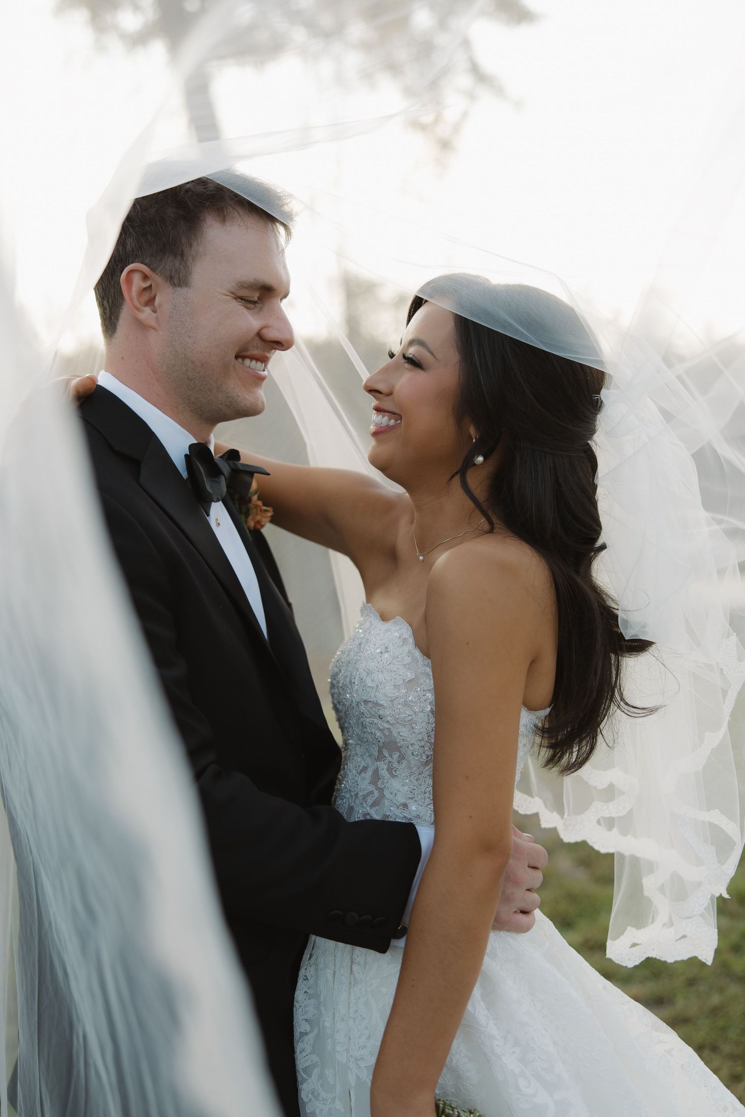 Smiling bride in a lace wedding gown and bridal veil embraces her groom in a black tuxedo and bow tie during an outdoor wedding portrait.