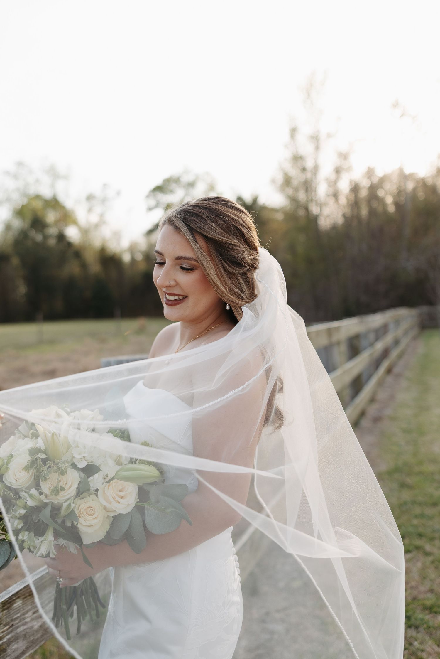 Bride in white gown and flowing veil holds white rose bouquet outdoors near a wooden fence at golden hour.