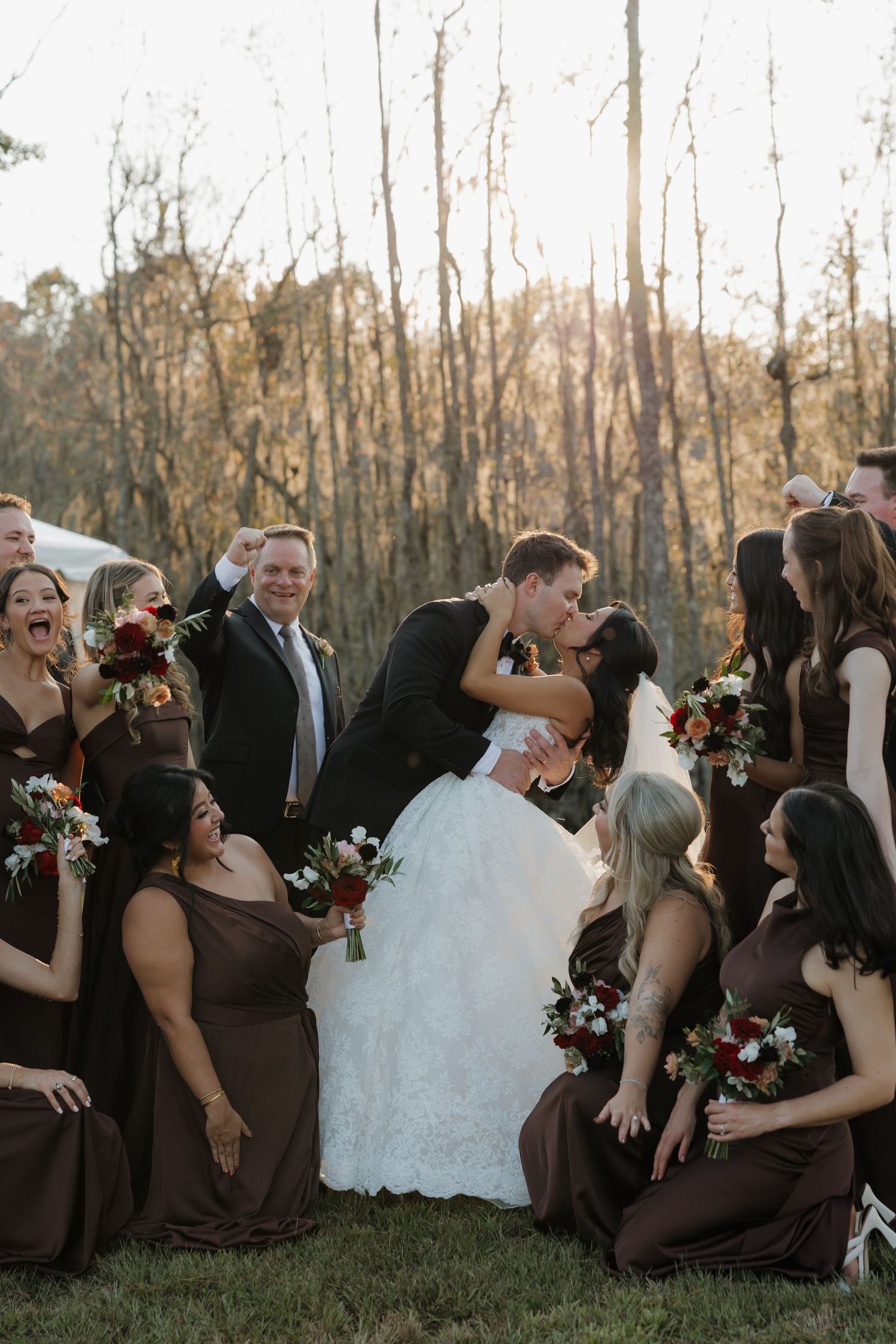 A bride and groom kiss while bridesmaids in brown dresses and guests laugh and cheer around them outdoors.
