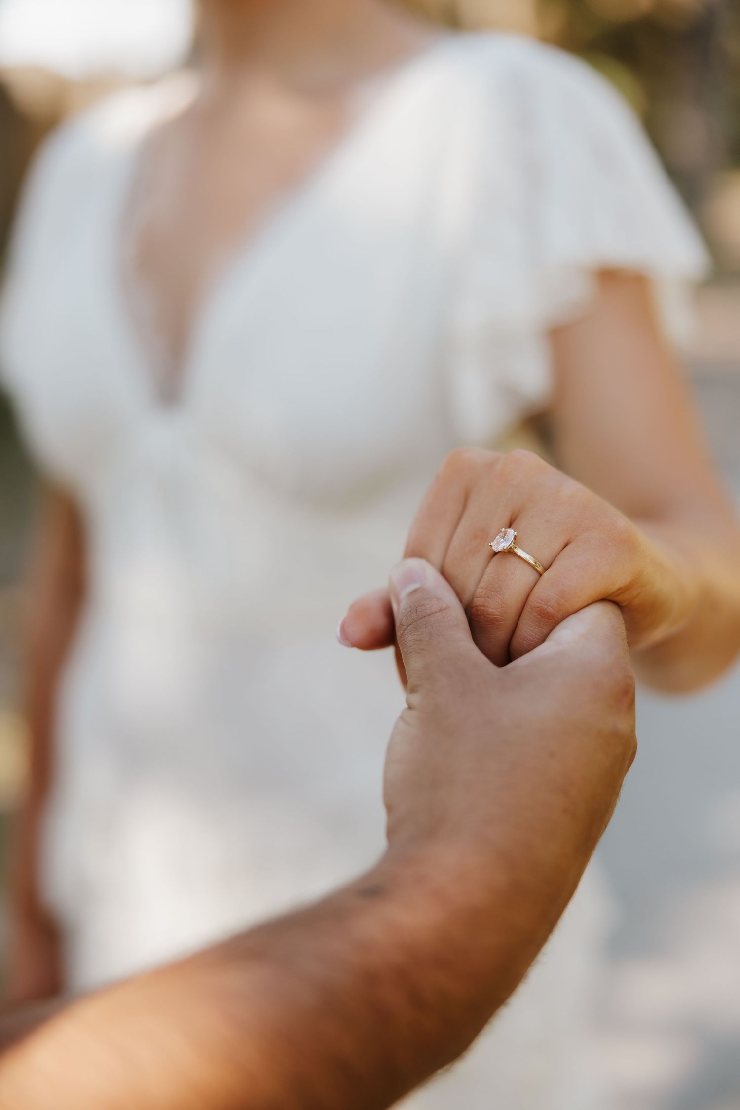 Close-up of a bride's hand wearing a gold marquise diamond engagement ring while holding her partner's hand.