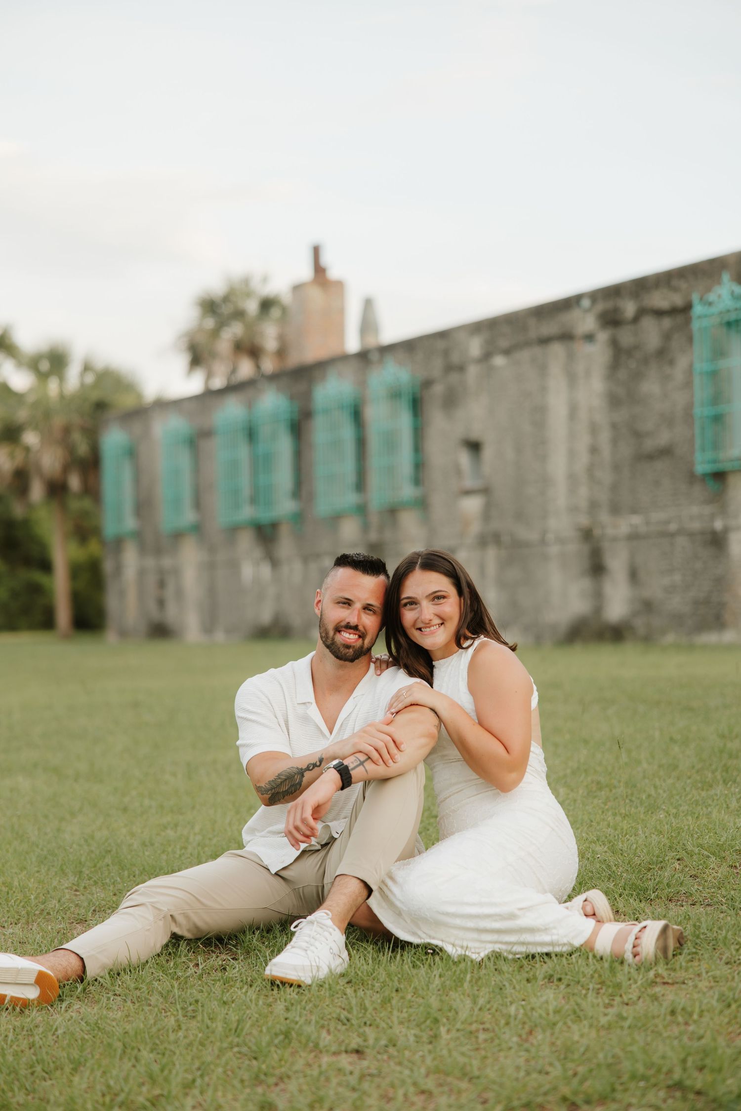 A smiling couple sits on green grass in front of a historic stone building with teal shutters.