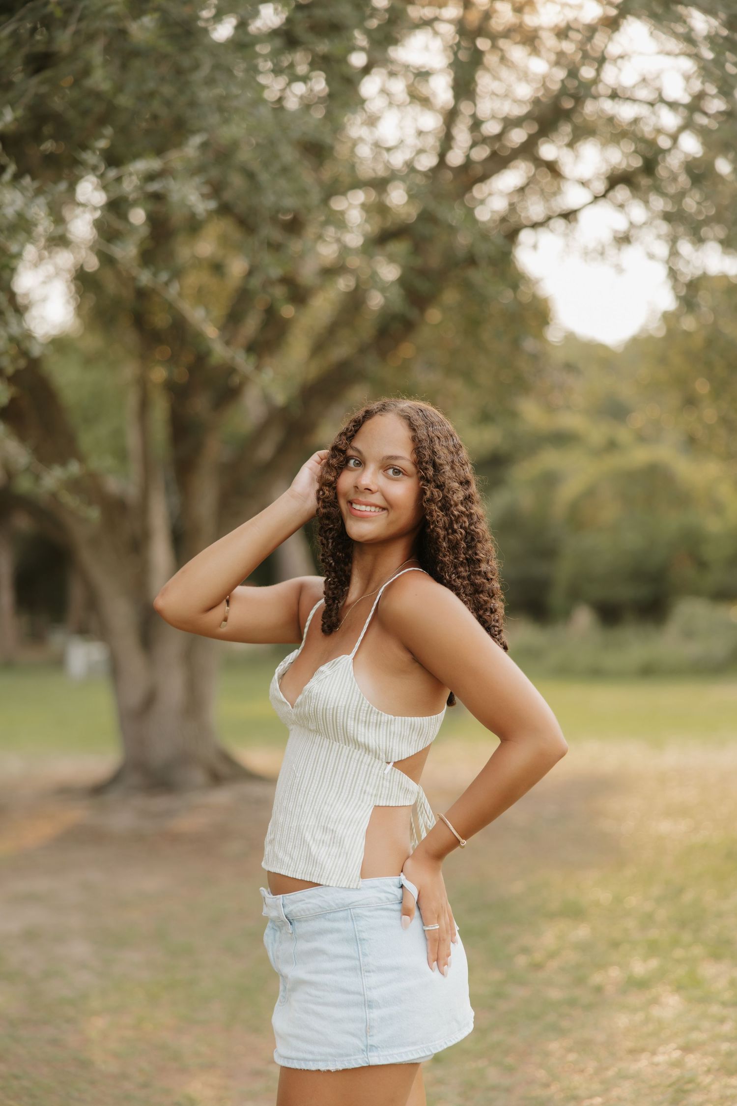 Young woman with curly hair in white crop top and denim mini skirt posing outdoors in a park setting.