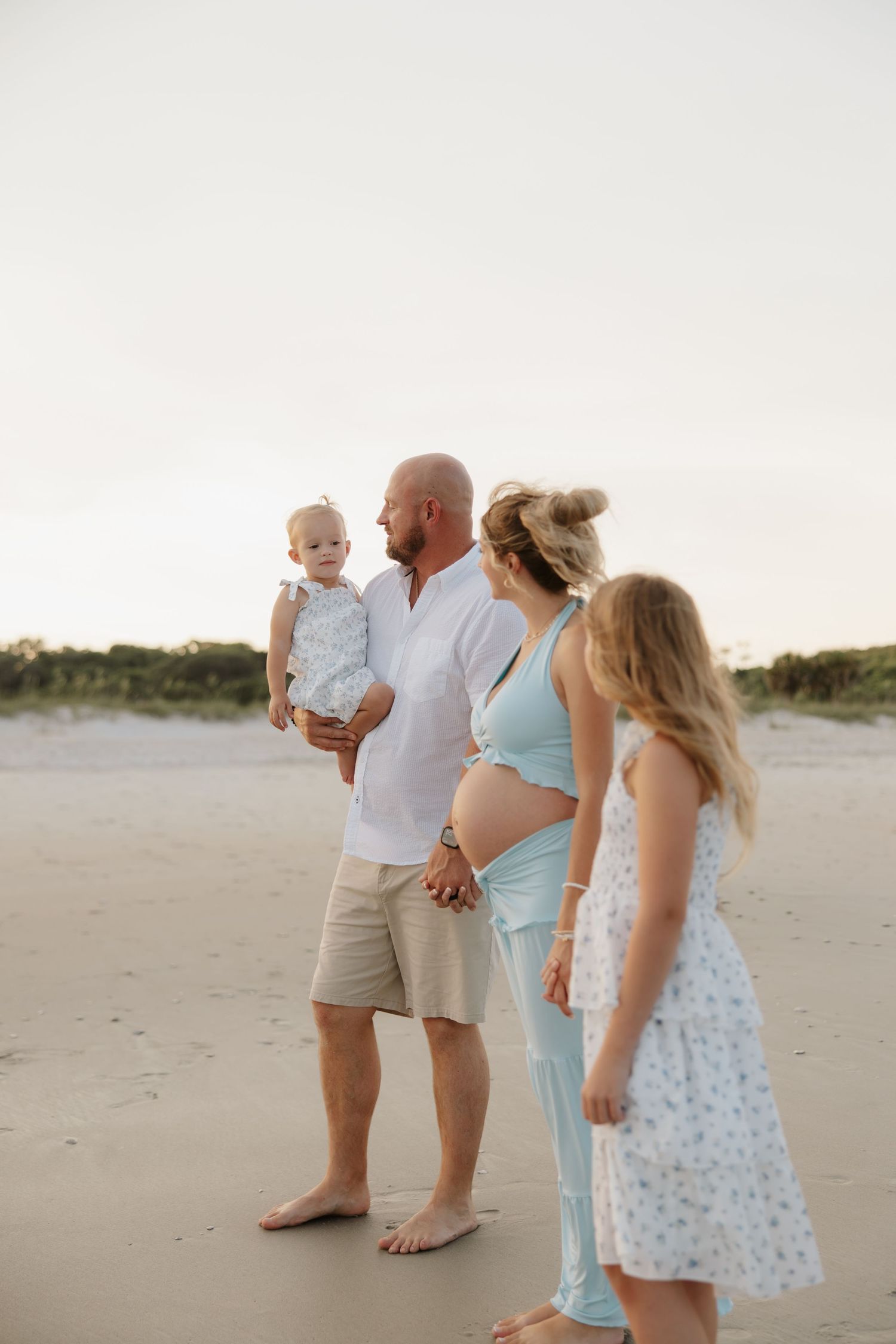 Pregnant family of four poses on sandy beach at sunset, father holding baby, older daughter nearby.