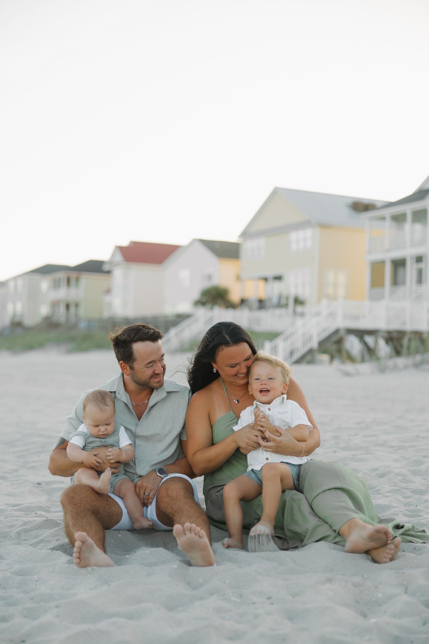 A happy family of four sits on a sandy beach, with two toddlers, beach houses visible in the background.