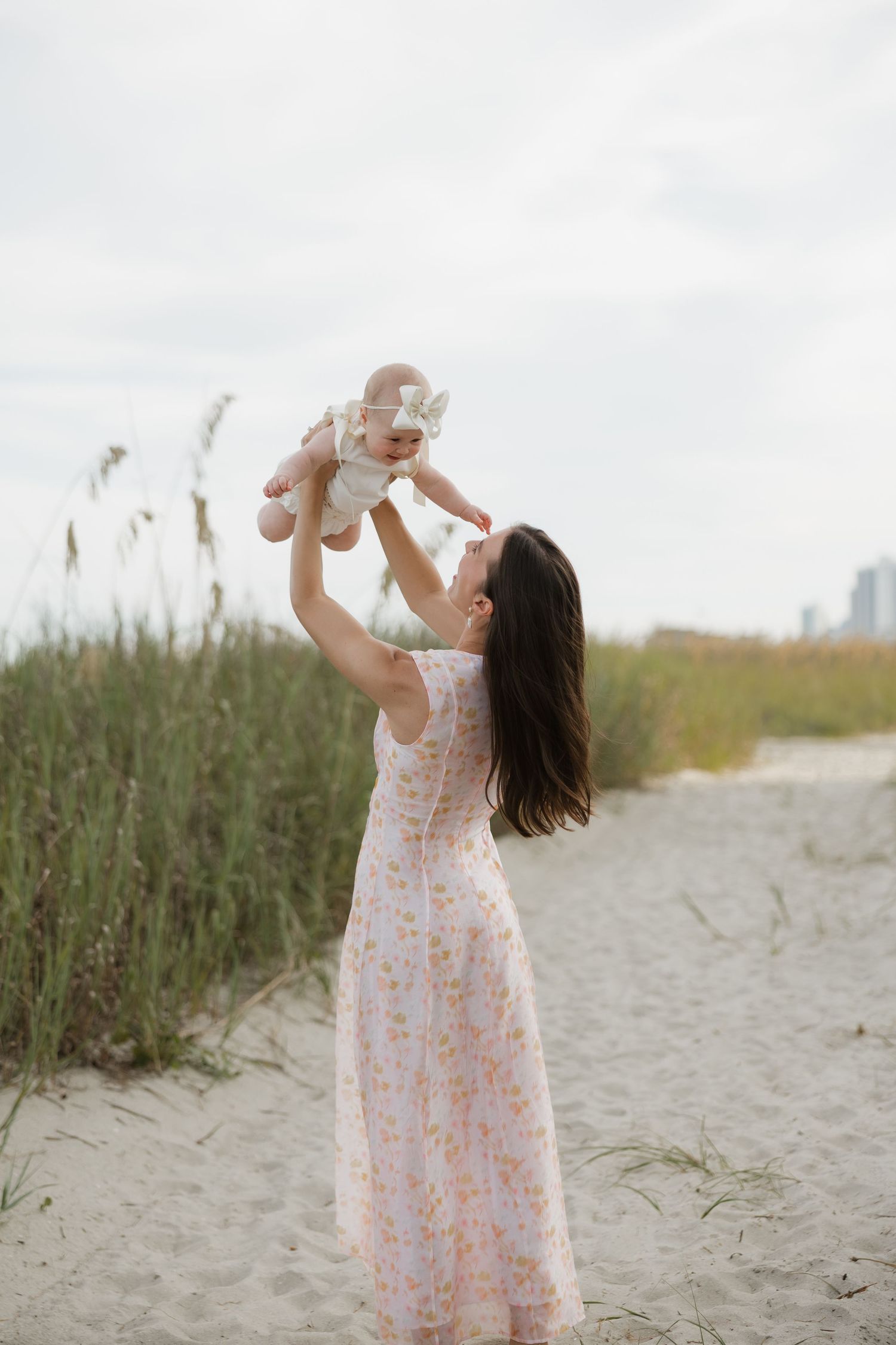 A woman in a floral dress lifts her baby joyfully on a sandy beach path surrounded by tall grass.