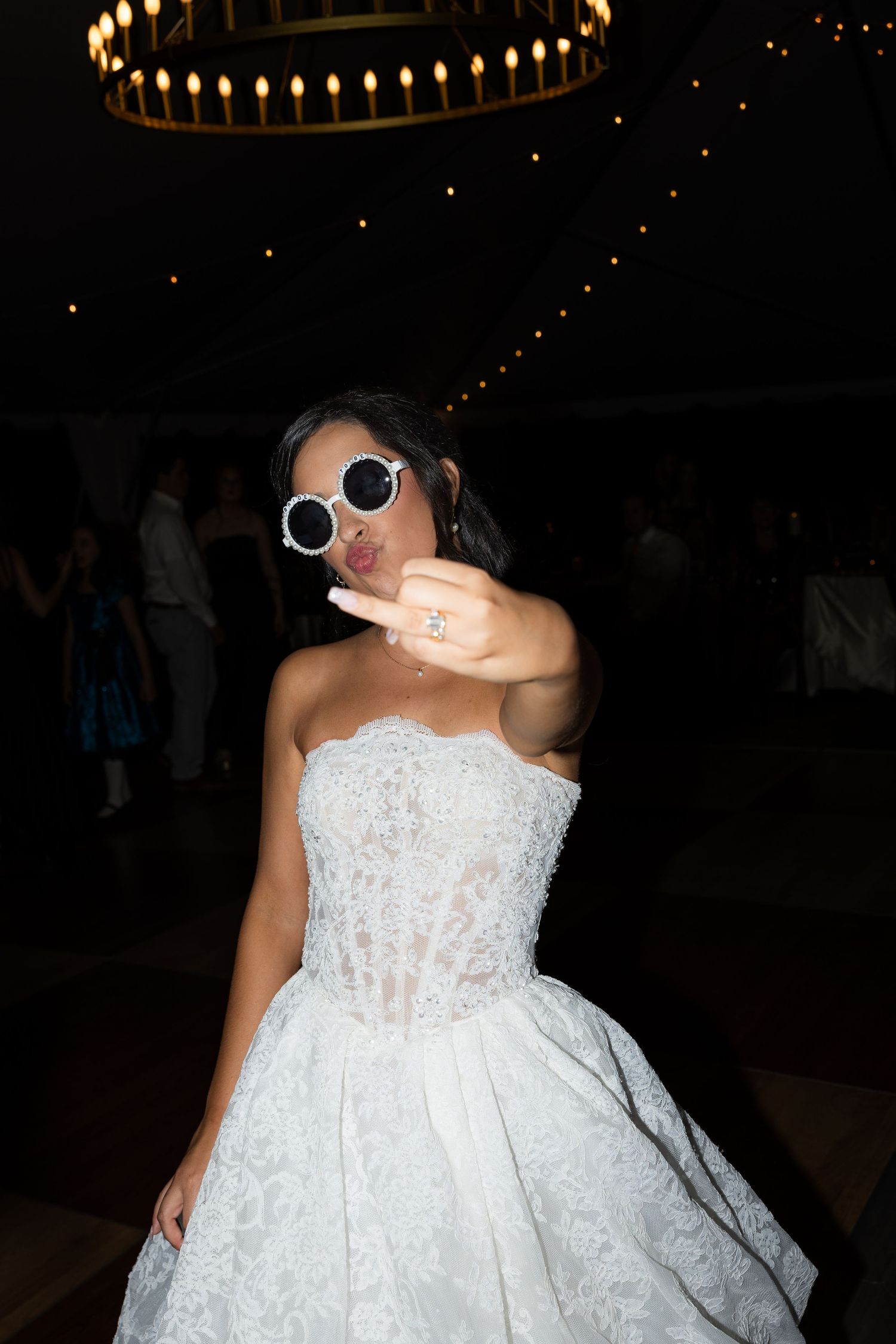 A bride in a lace strapless wedding gown and round white sunglasses blows a kiss and shows her ring on a dim dance floor.