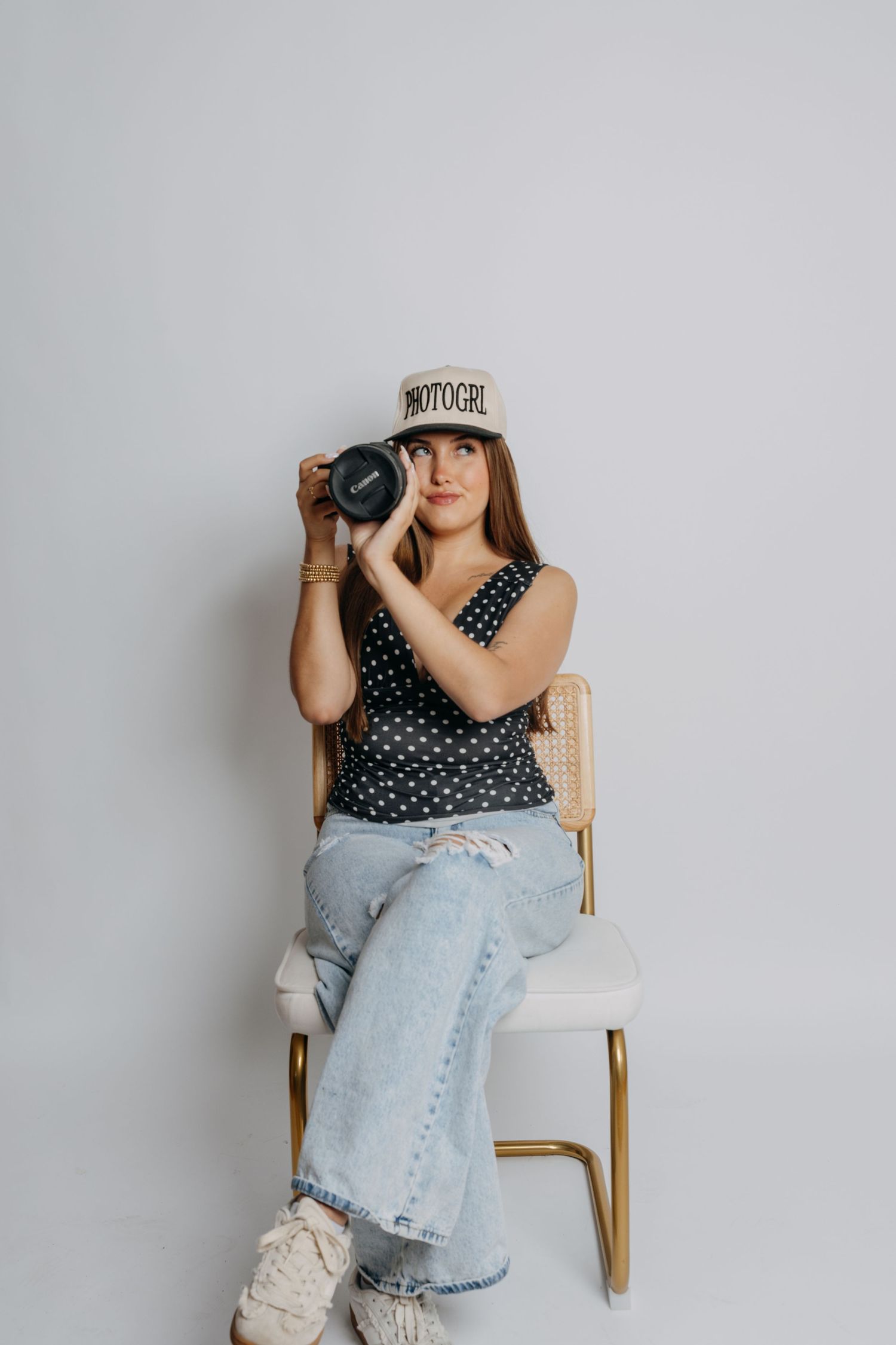 Woman in polka dot top and jeans sitting on chair holding DSLR camera wearing PHOTOGRL bucket hat.