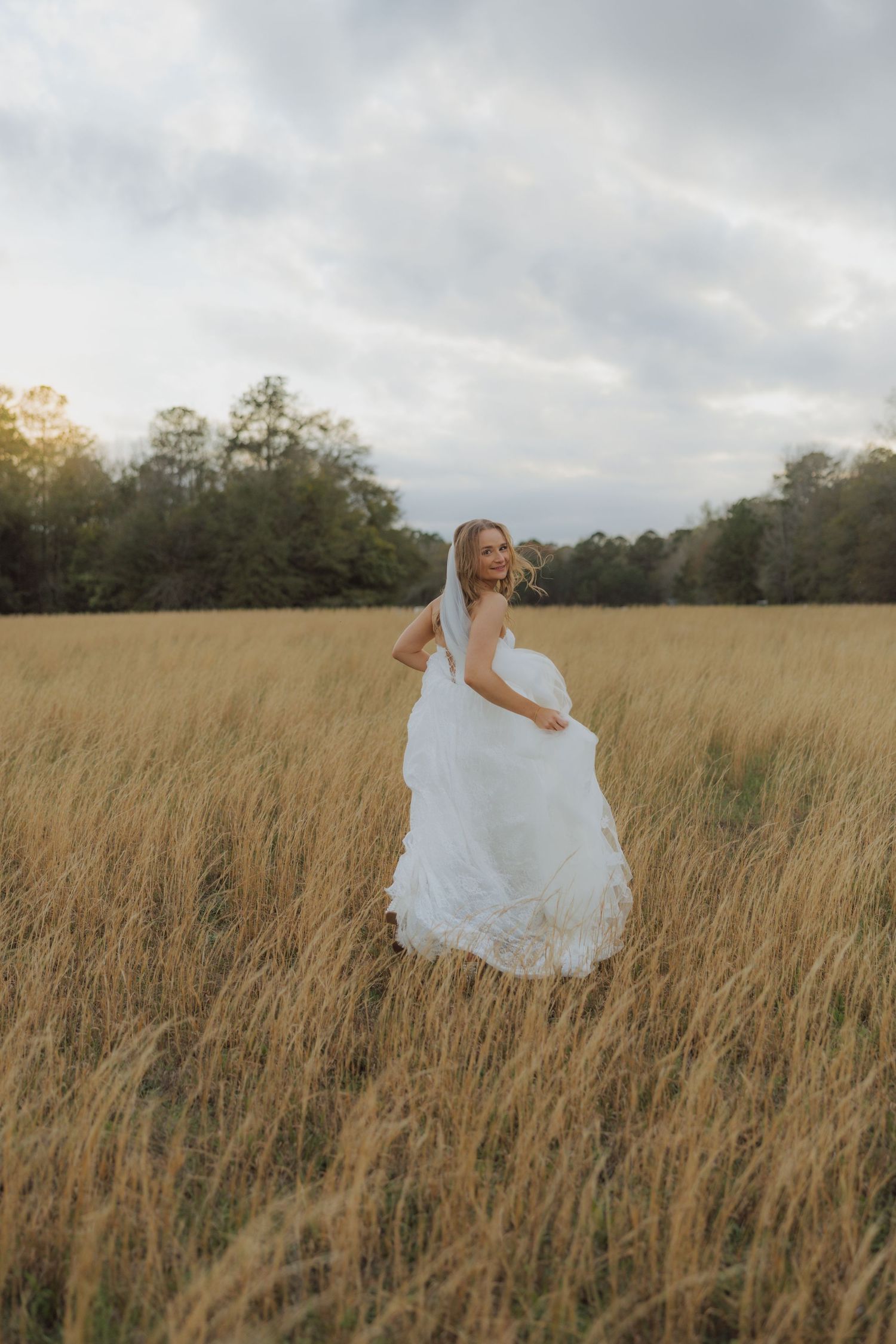 Bride in white lace gown and veil twirling in golden field at sunset.