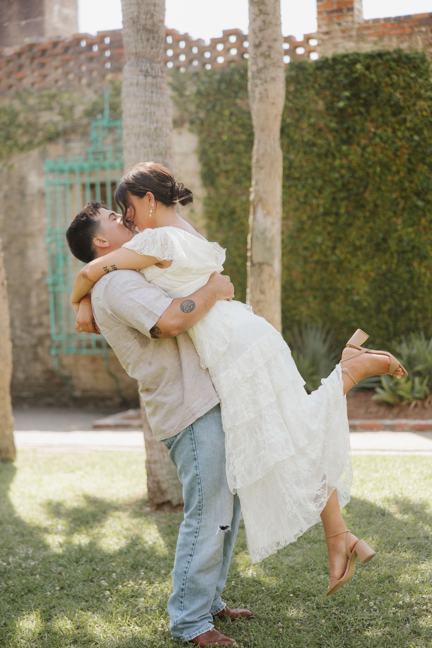 Couple sharing a romantic embrace outdoors, man lifting woman in white dress, surrounded by lush greenery.