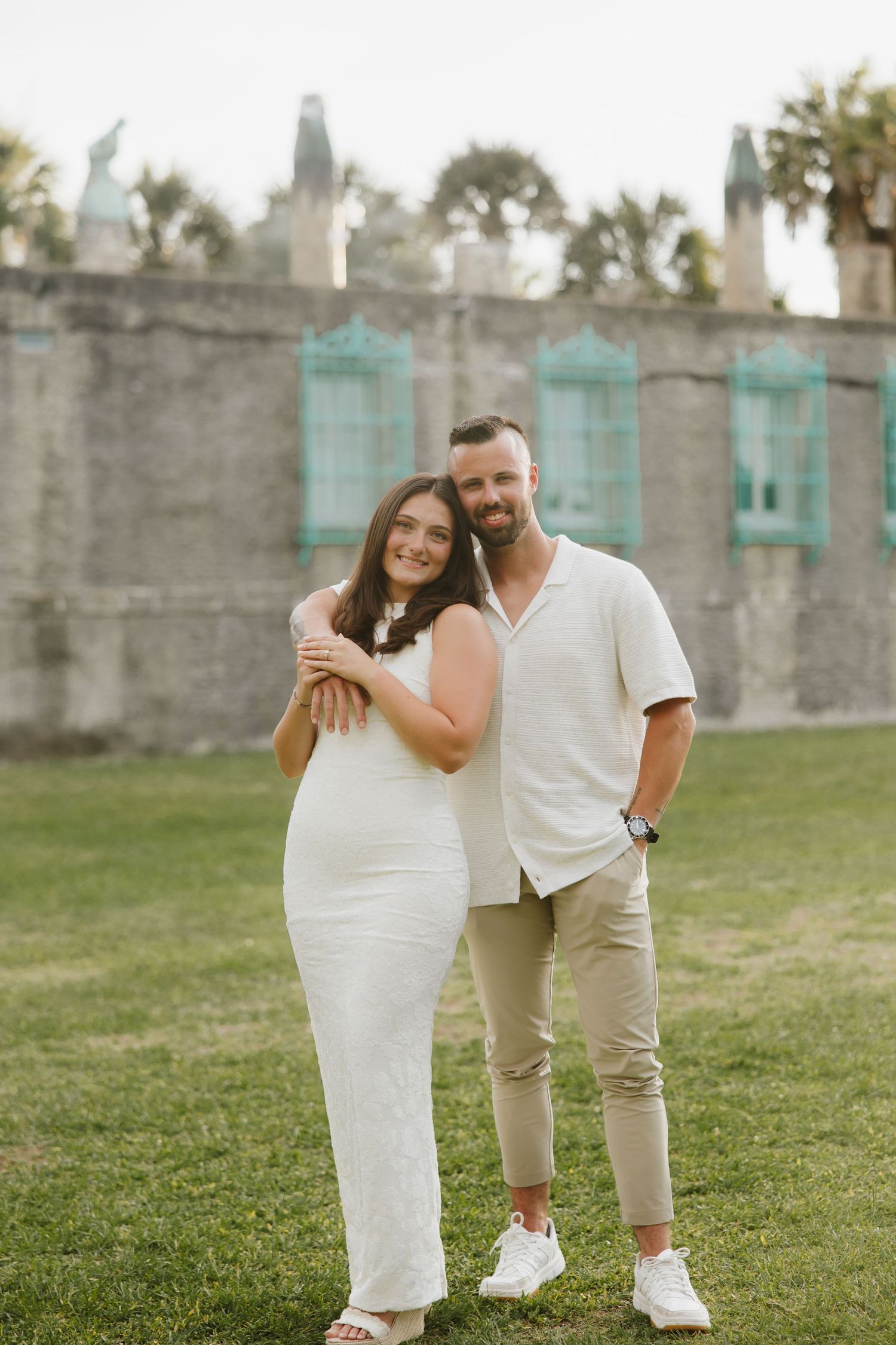 Smiling couple posing in front of historic stone building with turquoise windows on green lawn.