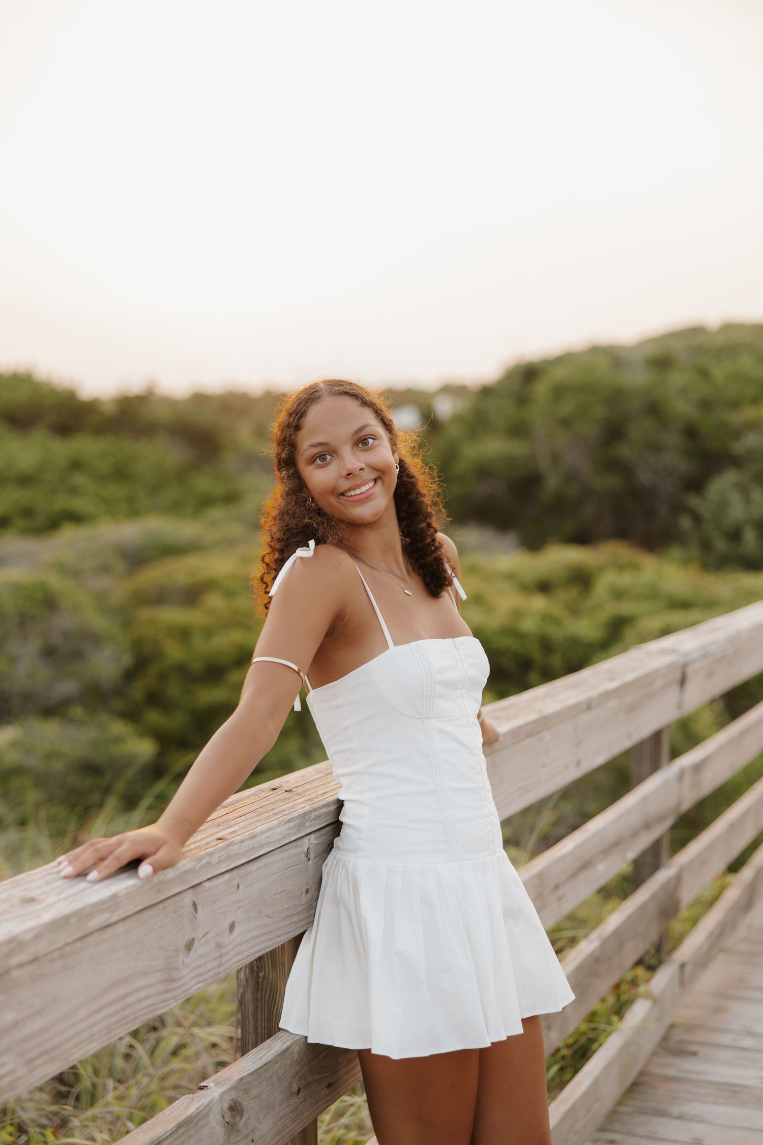 Young woman in white sundress smiling on wooden boardwalk at sunset with lush greenery behind her.