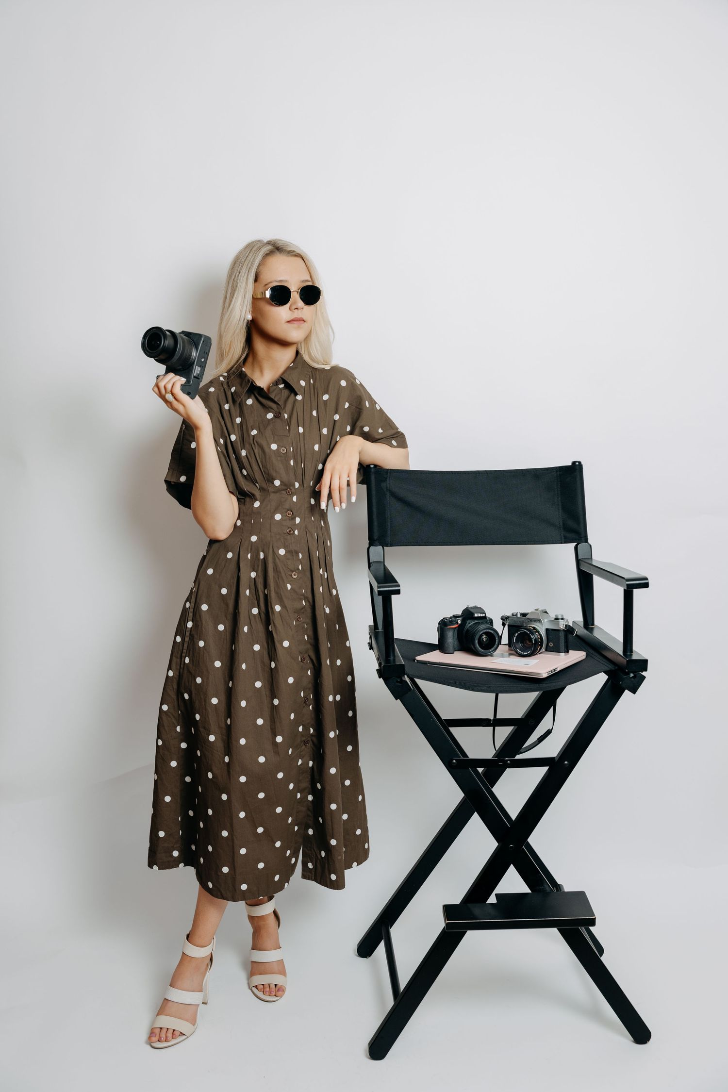 Blonde woman in polka dot dress holding camera beside director's chair with cameras.