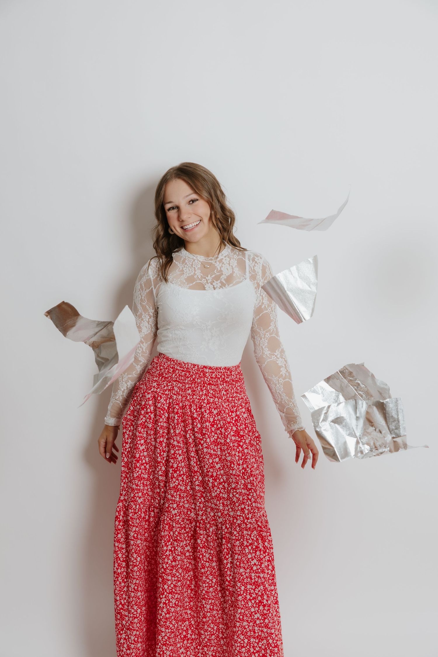 Smiling woman in lace top and floral skirt tosses silver foil highlights against a white wall.