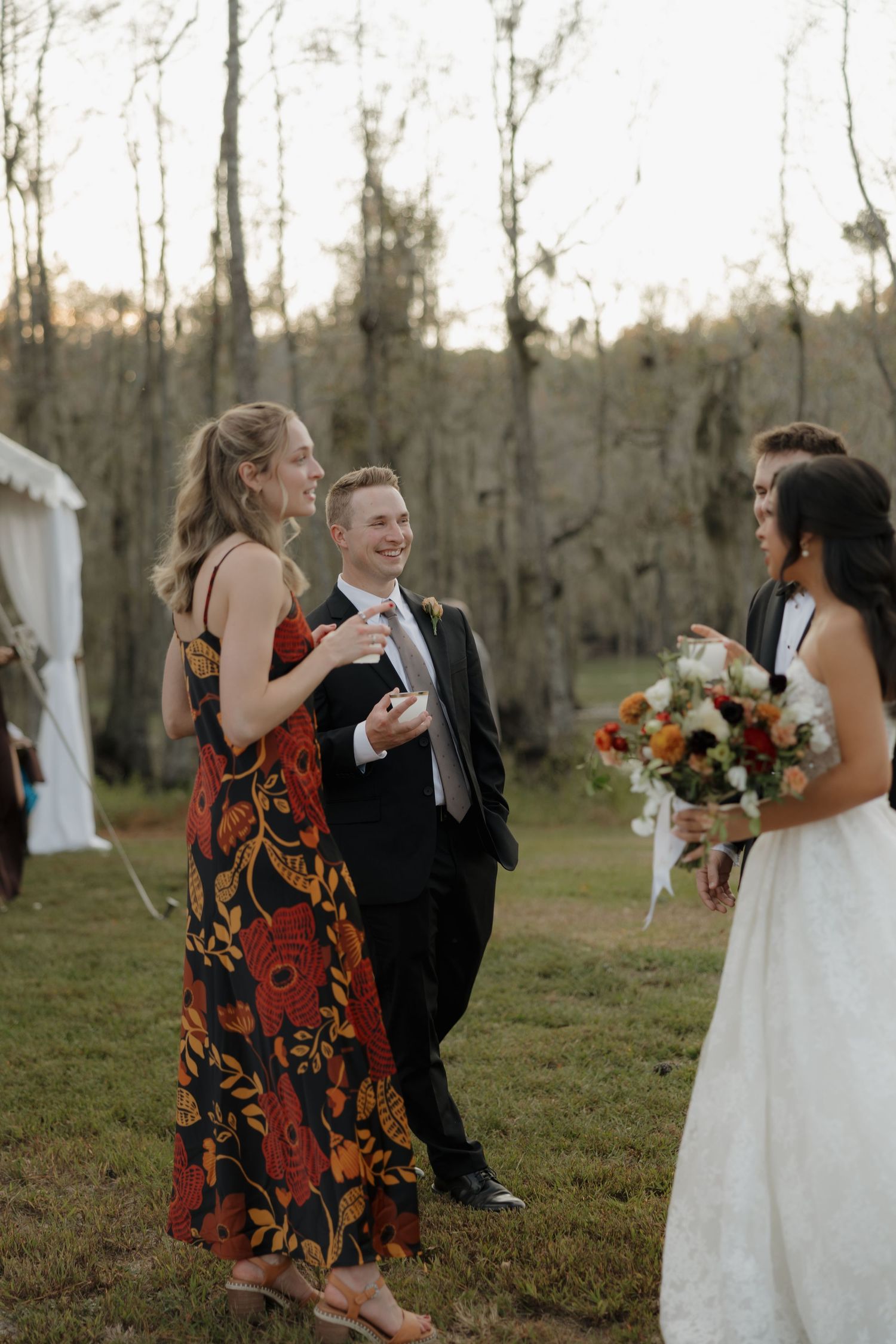 Wedding guests in a grassy field chat and laugh with a bride holding an autumn bouquet near a white tent and bare trees.