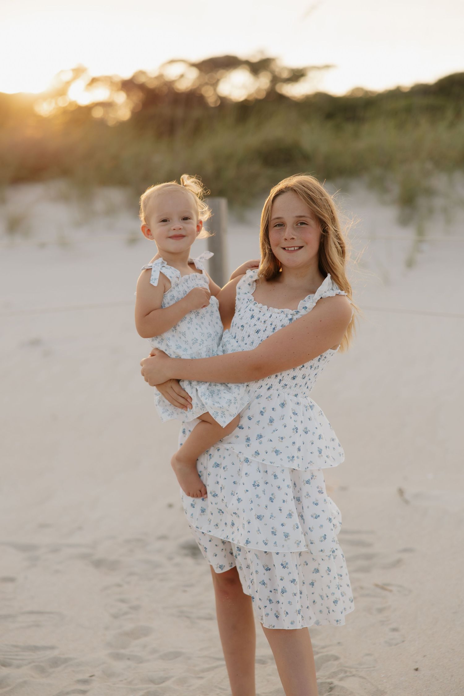 Older girl in white floral dress holds toddler sister on sandy beach at golden sunset.