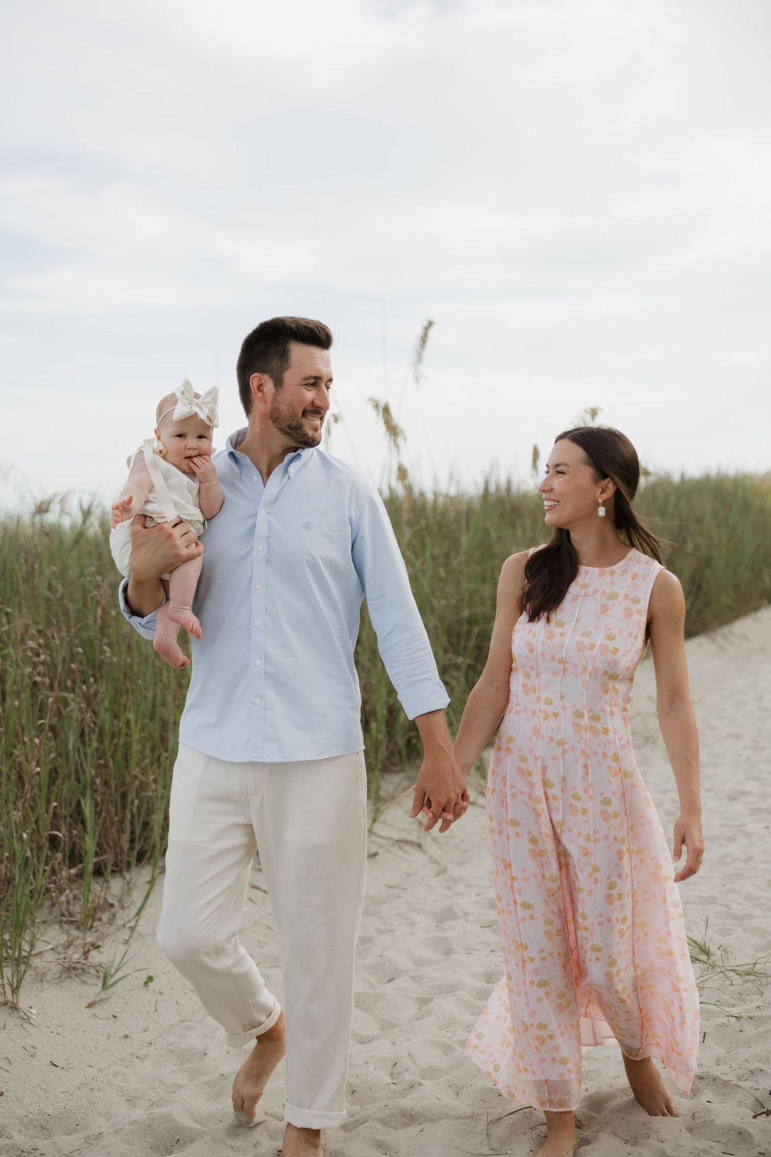 Happy young family walking on sandy beach, parents holding hands while father carries baby girl in floral outfit.