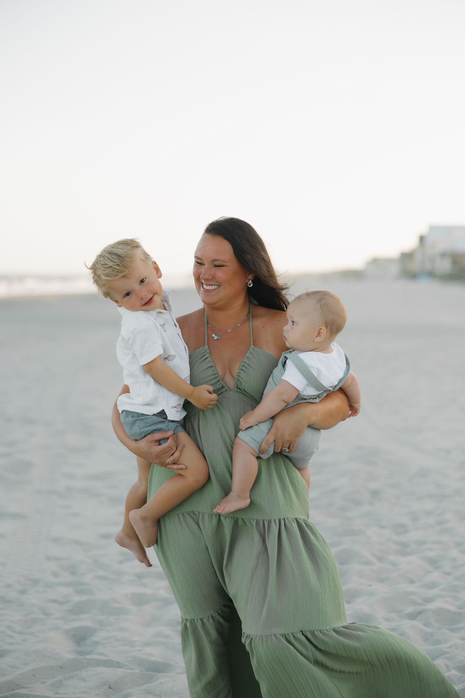 A smiling woman in a green maxi dress holds two young boys on a sandy beach at sunset.