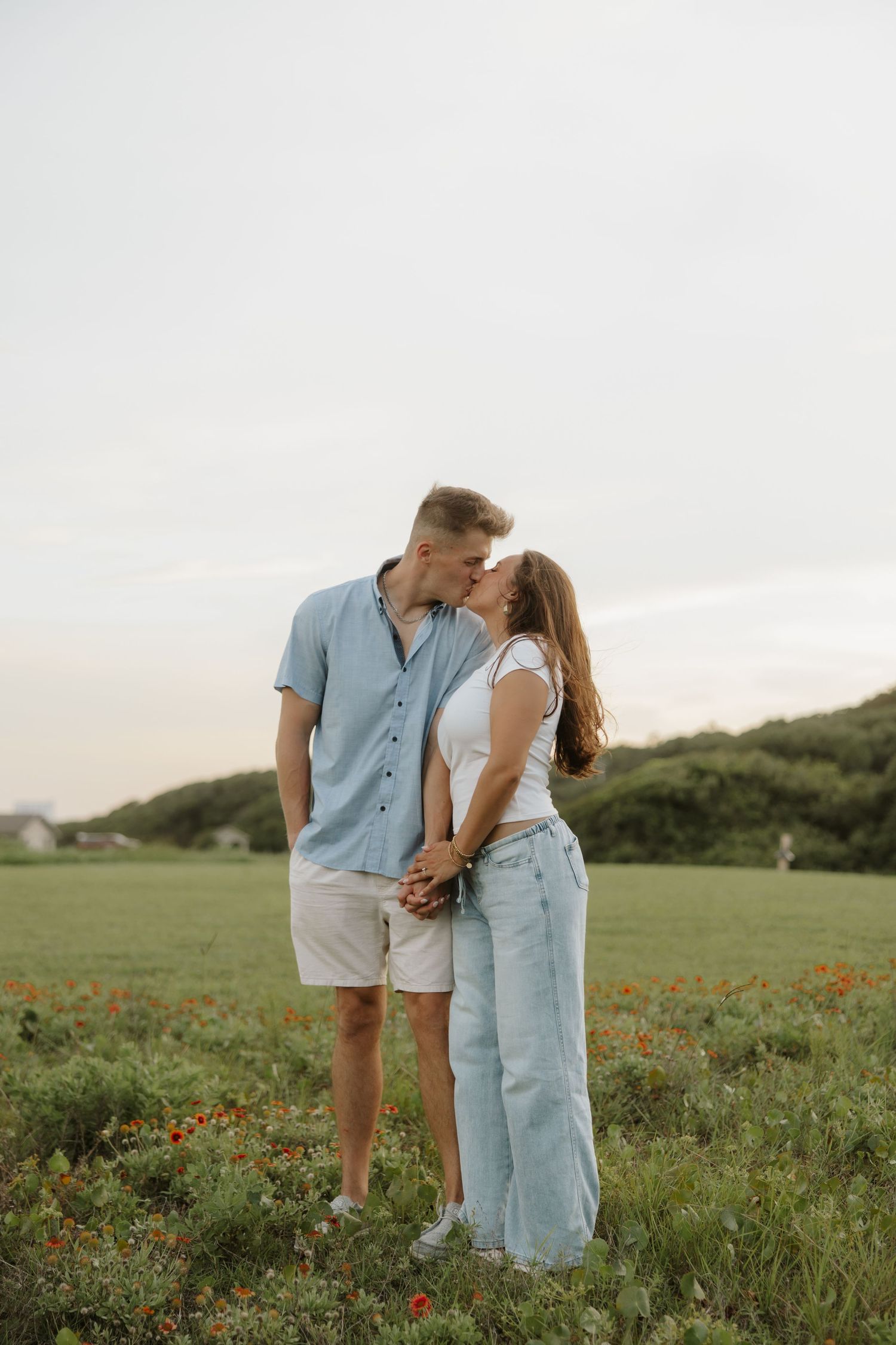Couple sharing a kiss in a green field with wildflowers, wearing casual summer outfits under a soft cloudy sky.