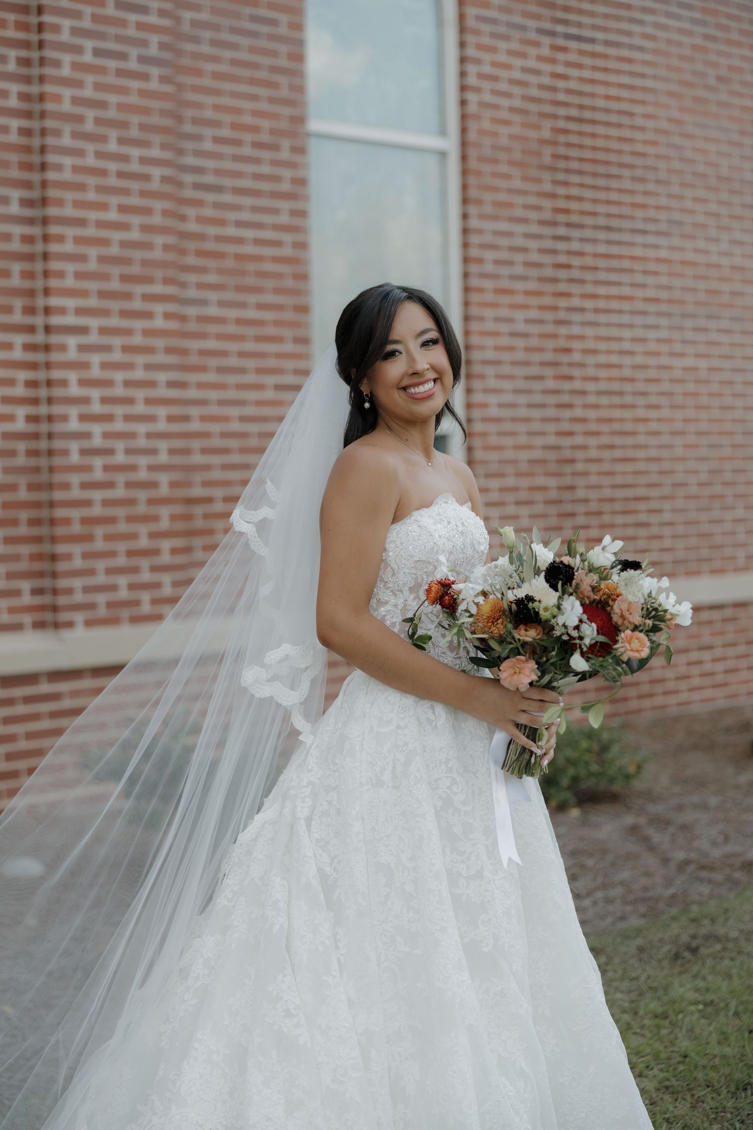 Smiling bride in a strapless lace gown and long veil holds a colorful floral bouquet in front of a red brick building.