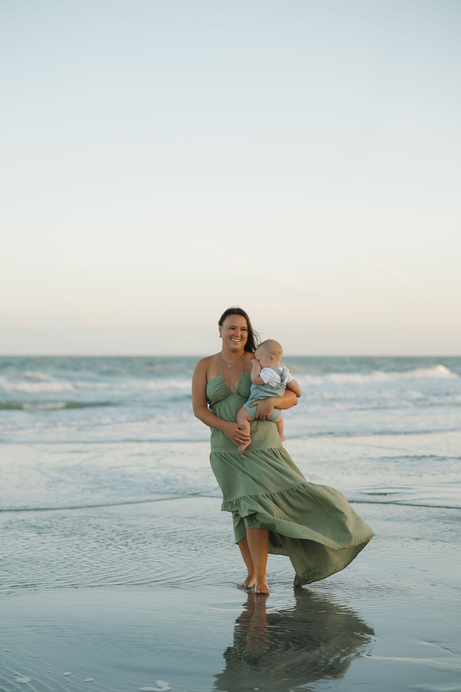 A smiling mother in a flowing green dress holds her baby while walking along the beach at sunset.