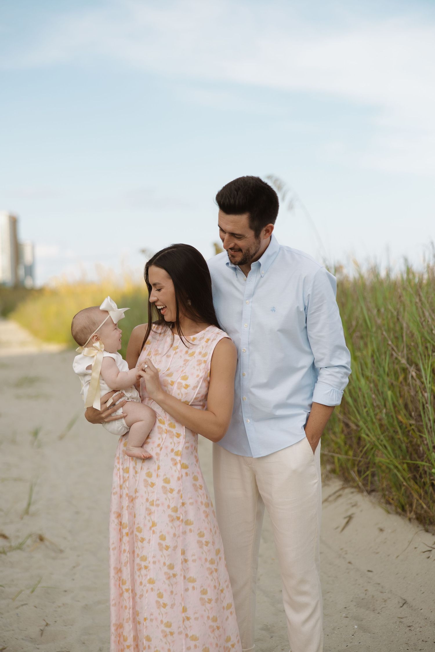 Young family with infant baby girl posing on sandy beach path surrounded by sea grass.