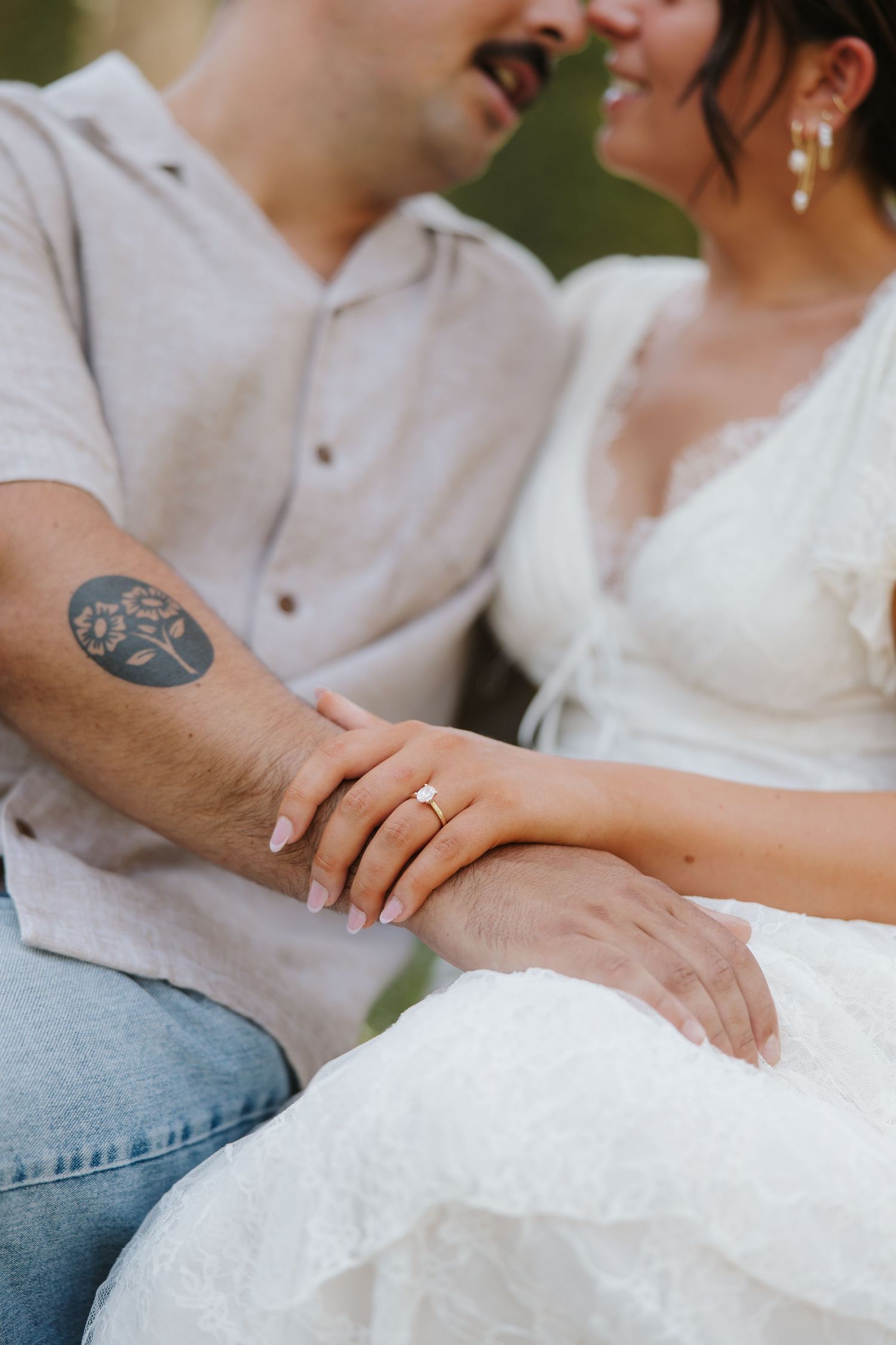 Couple holding hands showing engagement ring, man with floral tattoo wearing linen shirt, woman in white lace dress.