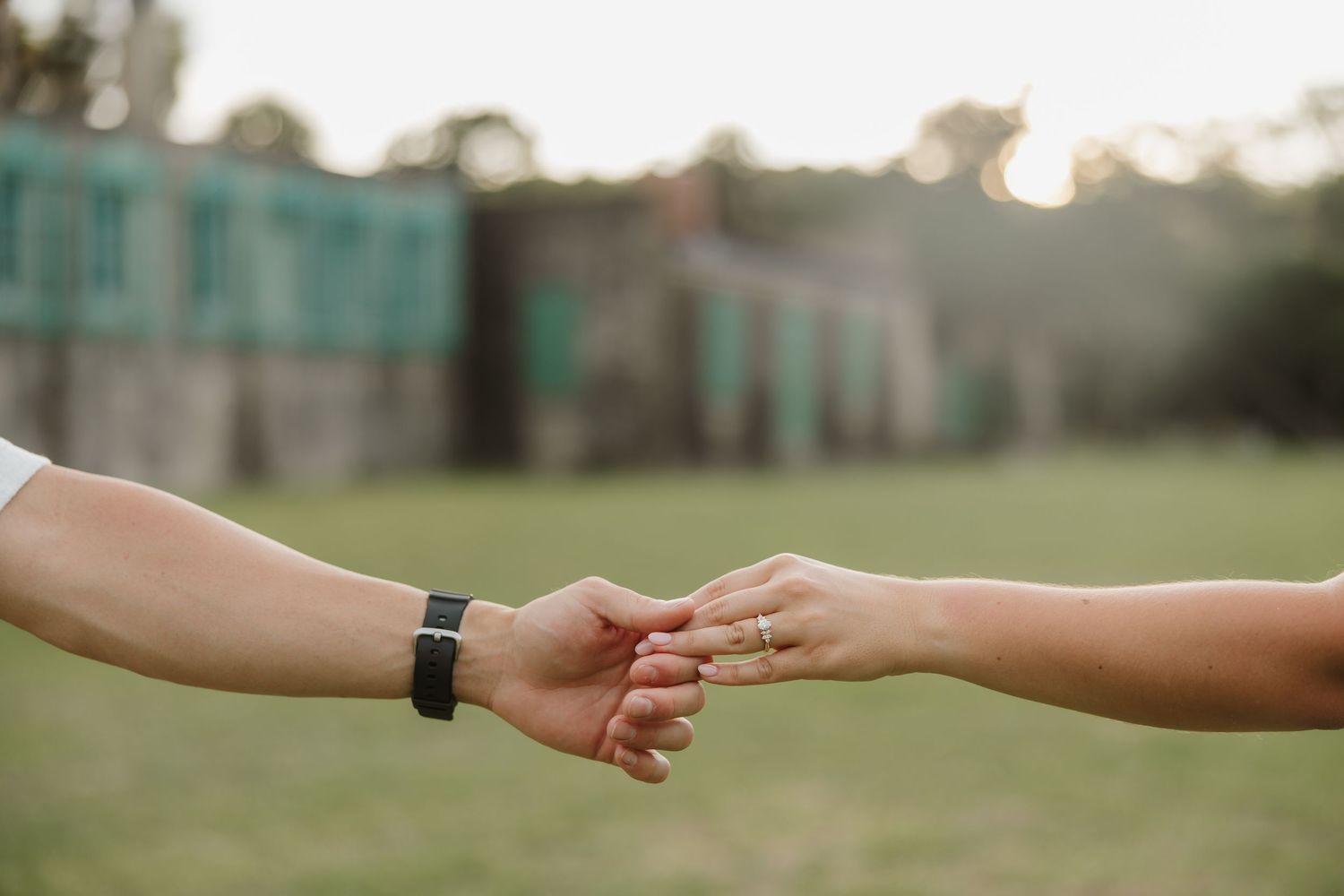 Two people gently holding hands outdoors with a blurred green building and lawn in the background.