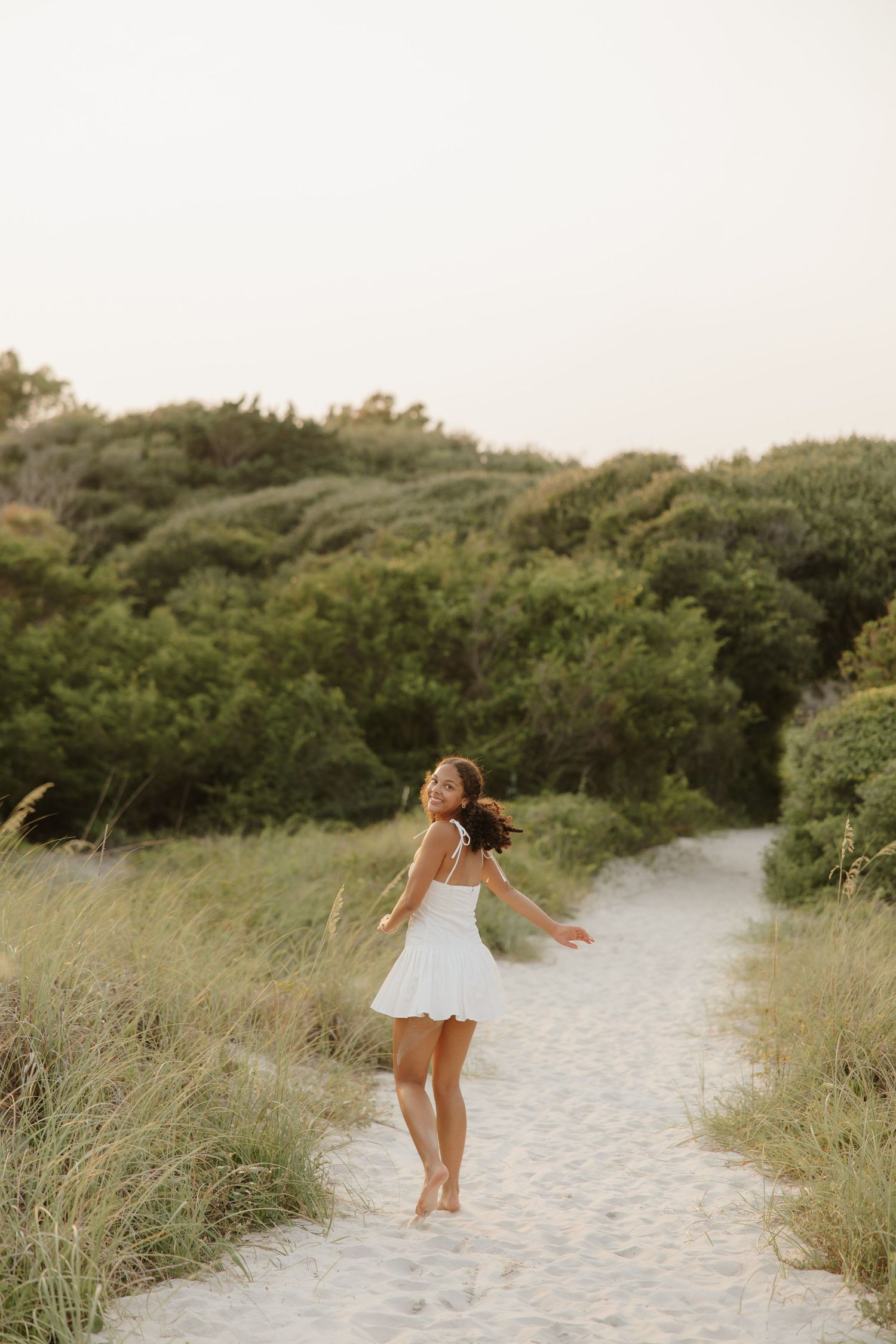 Young woman in white dress walking barefoot on sandy beach path surrounded by lush green dunes.