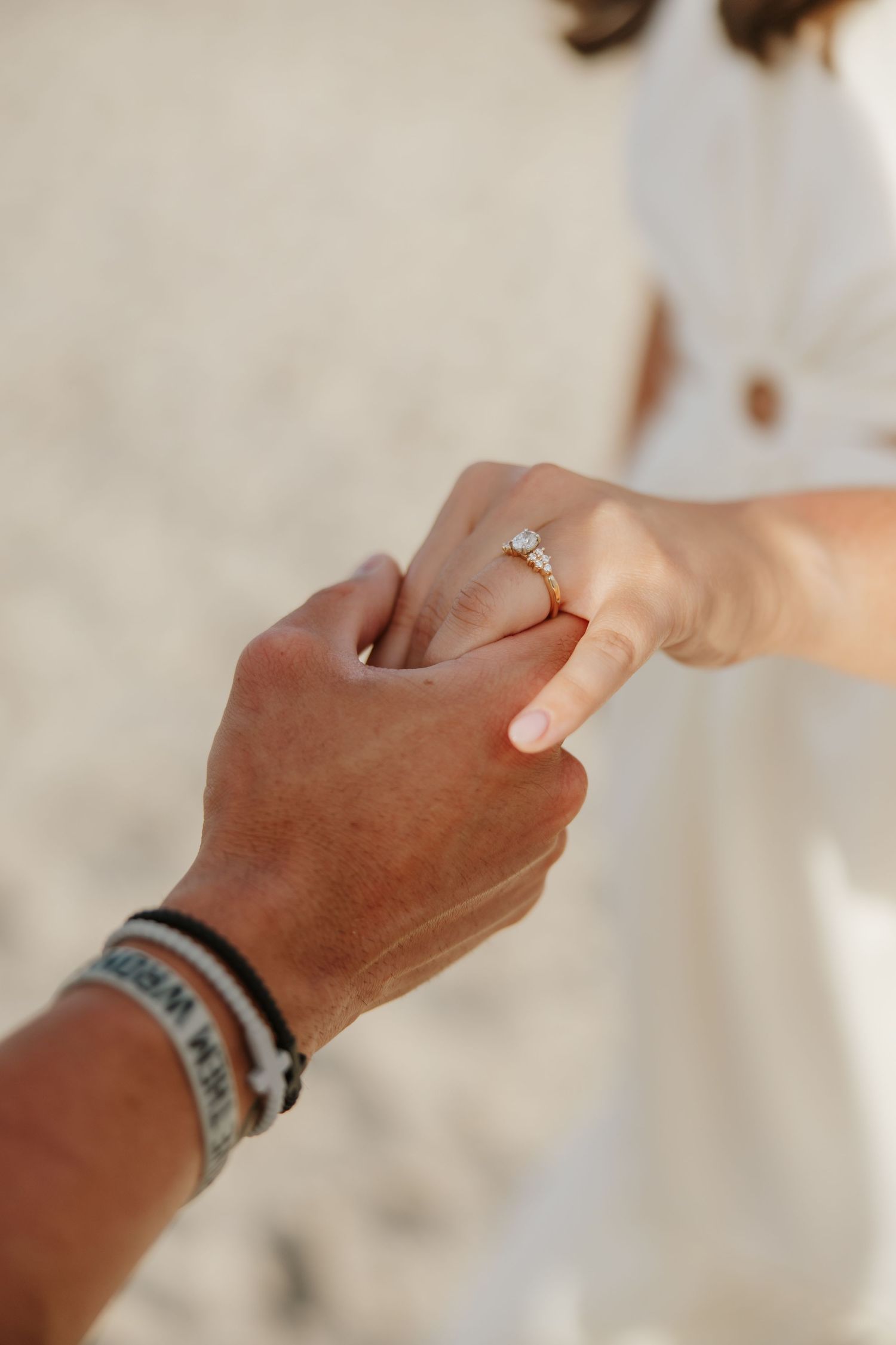 Close-up of couple holding hands on beach, woman wearing elegant diamond engagement ring, man with wristbands.