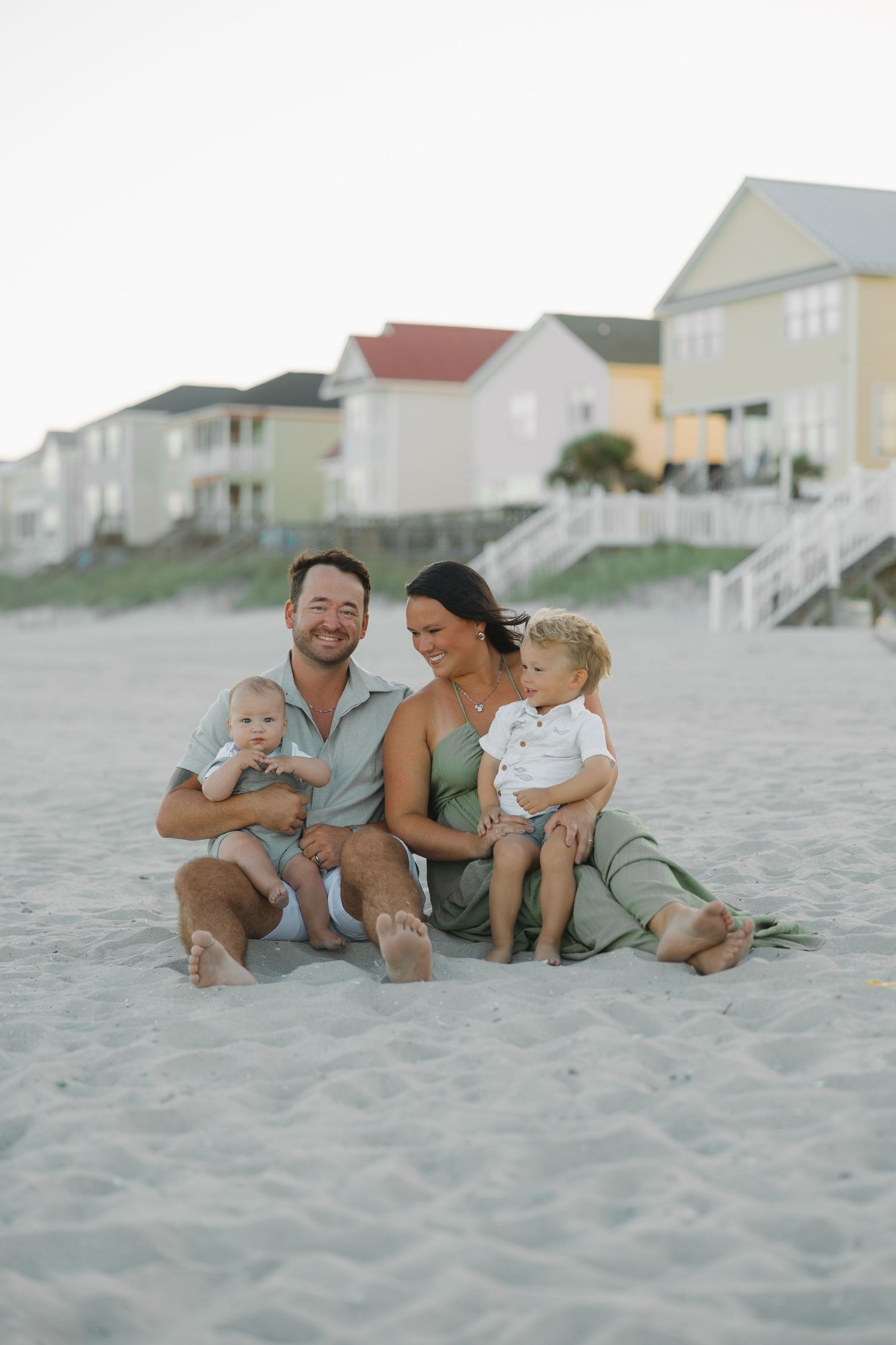 A happy family of four sits barefoot on a sandy beach, with colorful beach houses in the background.