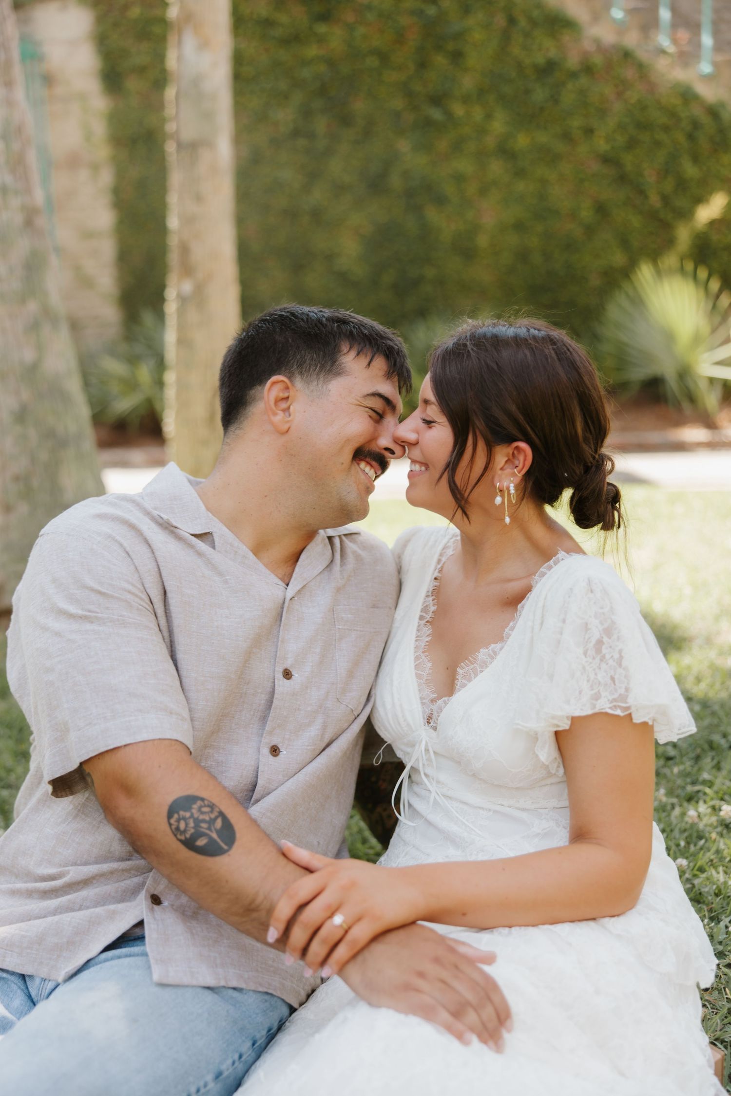 A couple shares a romantic nose-to-nose moment outdoors, the woman in white lace dress and man in beige shirt with arm tattoo.