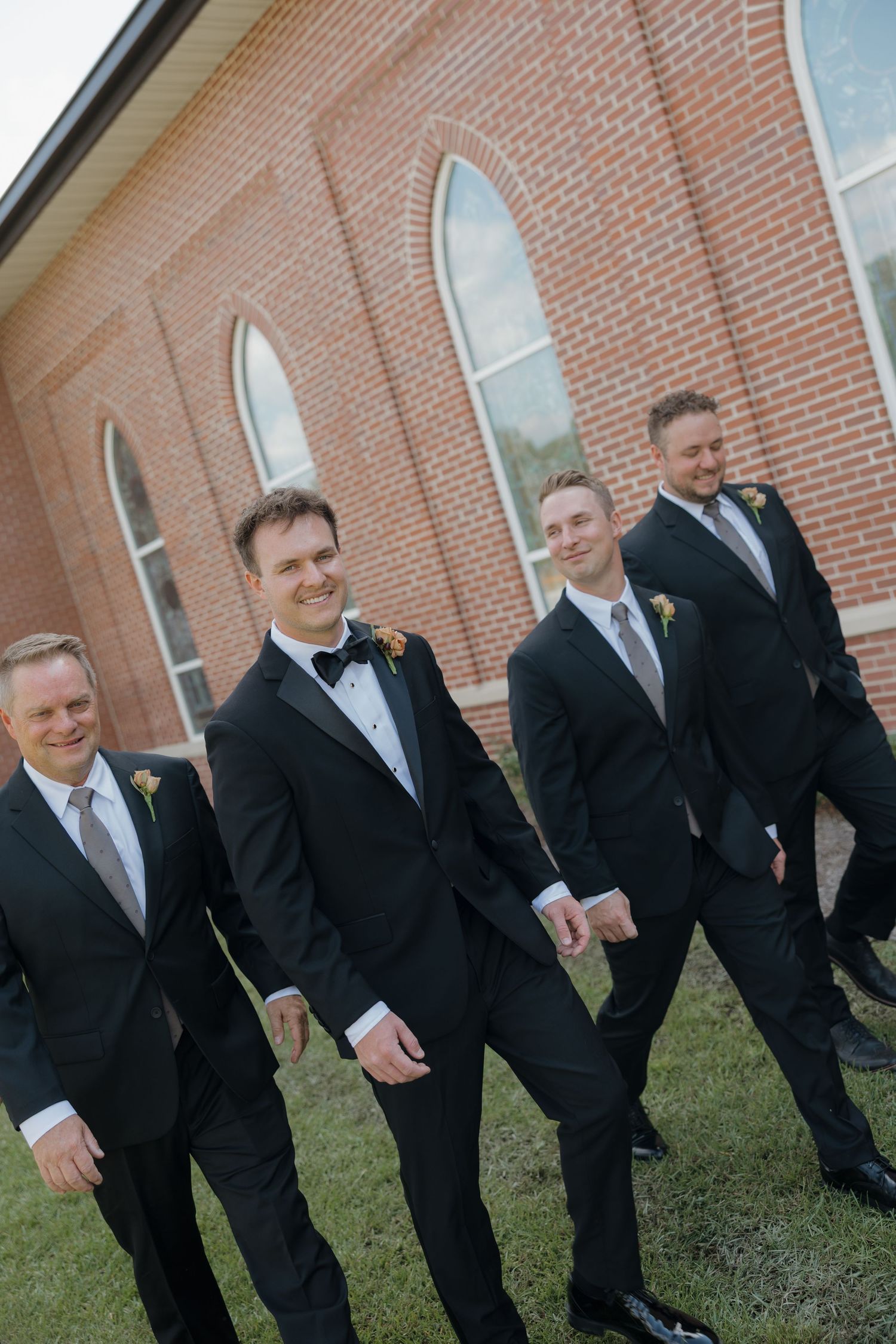 Four groomsmen in black tuxedos and boutonnieres walk and smile together on the lawn beside a brick building.