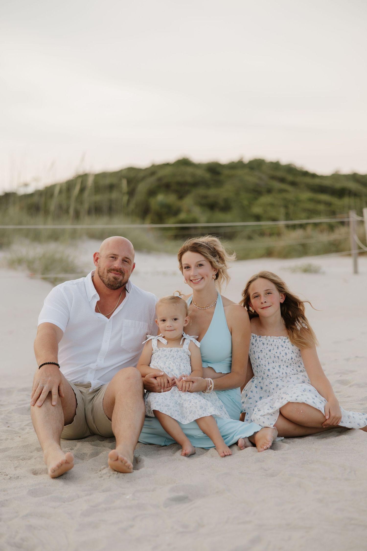 Happy family of four sits together on sandy beach at dusk in coordinated outfits.