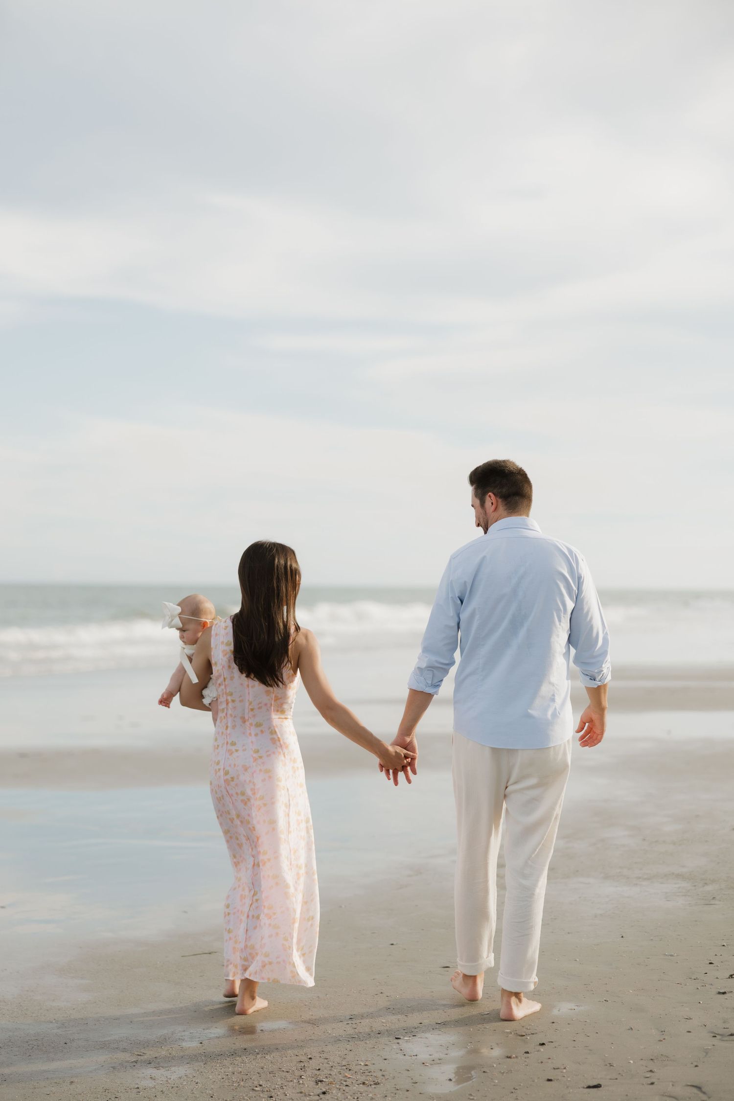 A young family walks hand-in-hand on a beach, with a mother carrying a baby and father beside her.