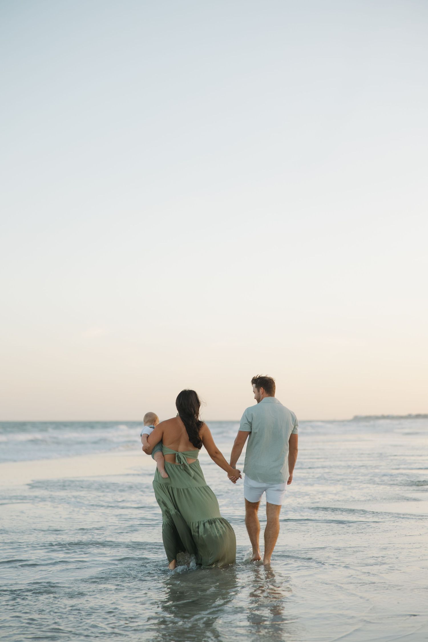 A couple holding hands walks along the ocean shore at sunset, with the woman carrying a baby in a flowing green dress.