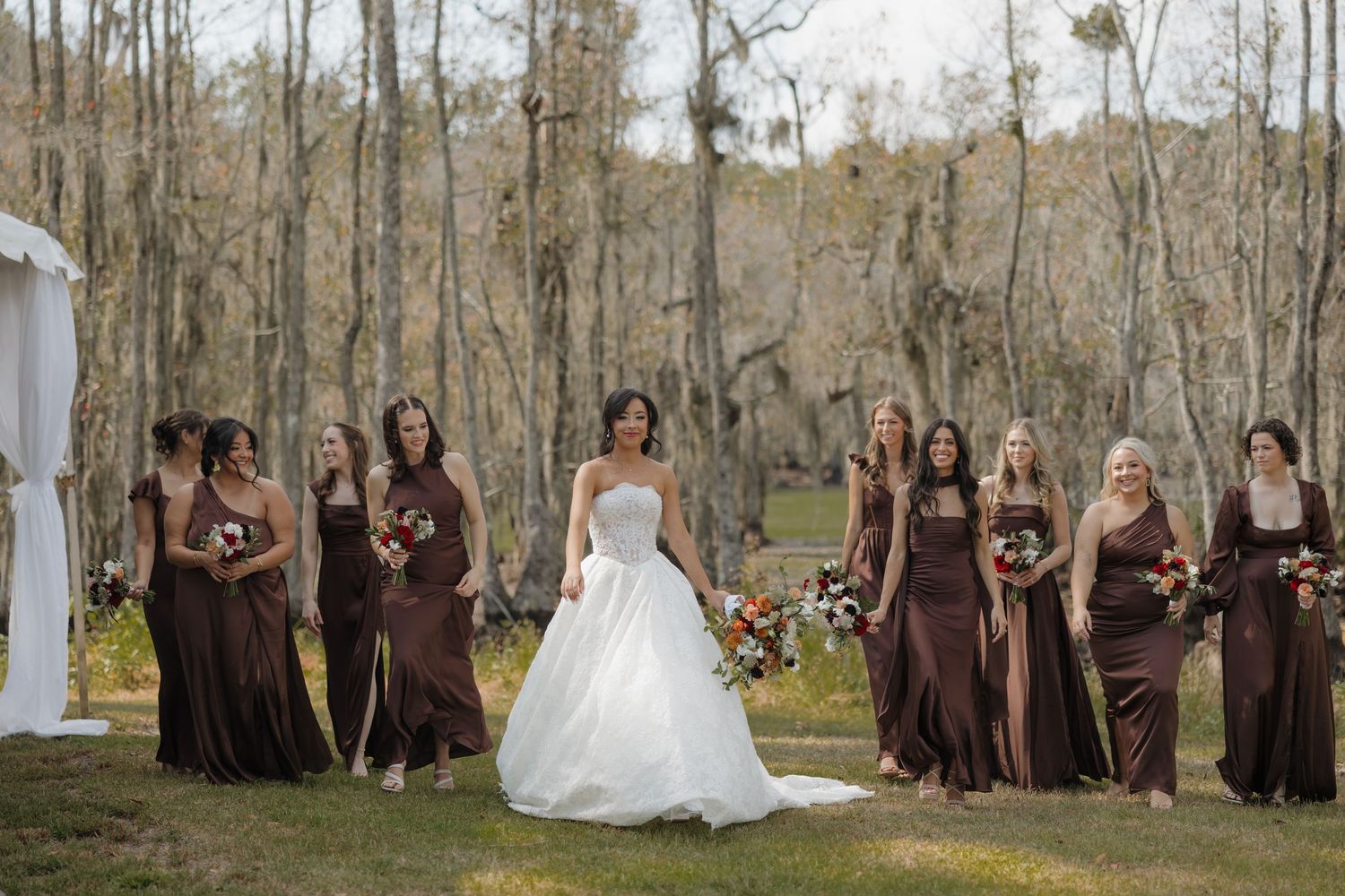 Bride in a white gown stands among bridesmaids in brown dresses holding bouquets on a grassy lawn surrounded by tall trees.