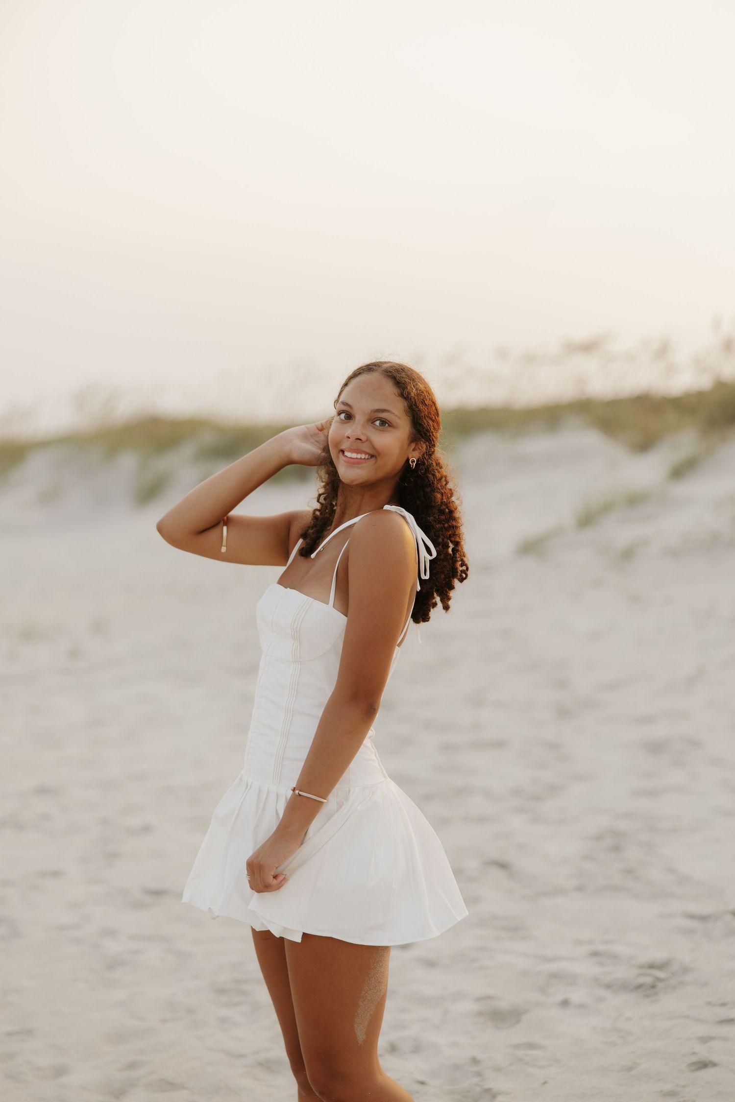 Smiling young woman in white mini dress posing on beach at golden hour with sand dunes behind her.