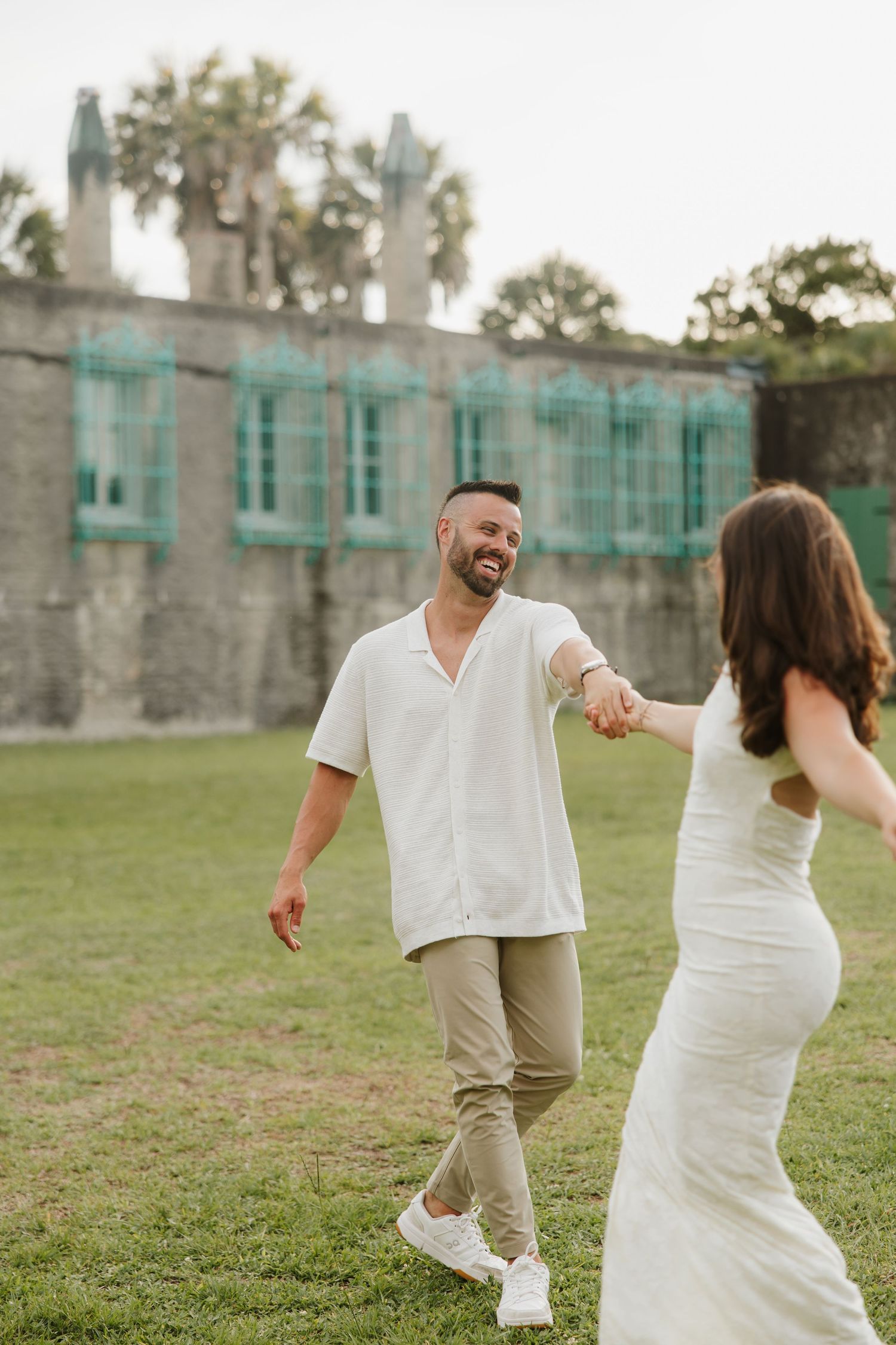 Smiling man in white shirt holds hands with woman in white dress walking on green lawn.