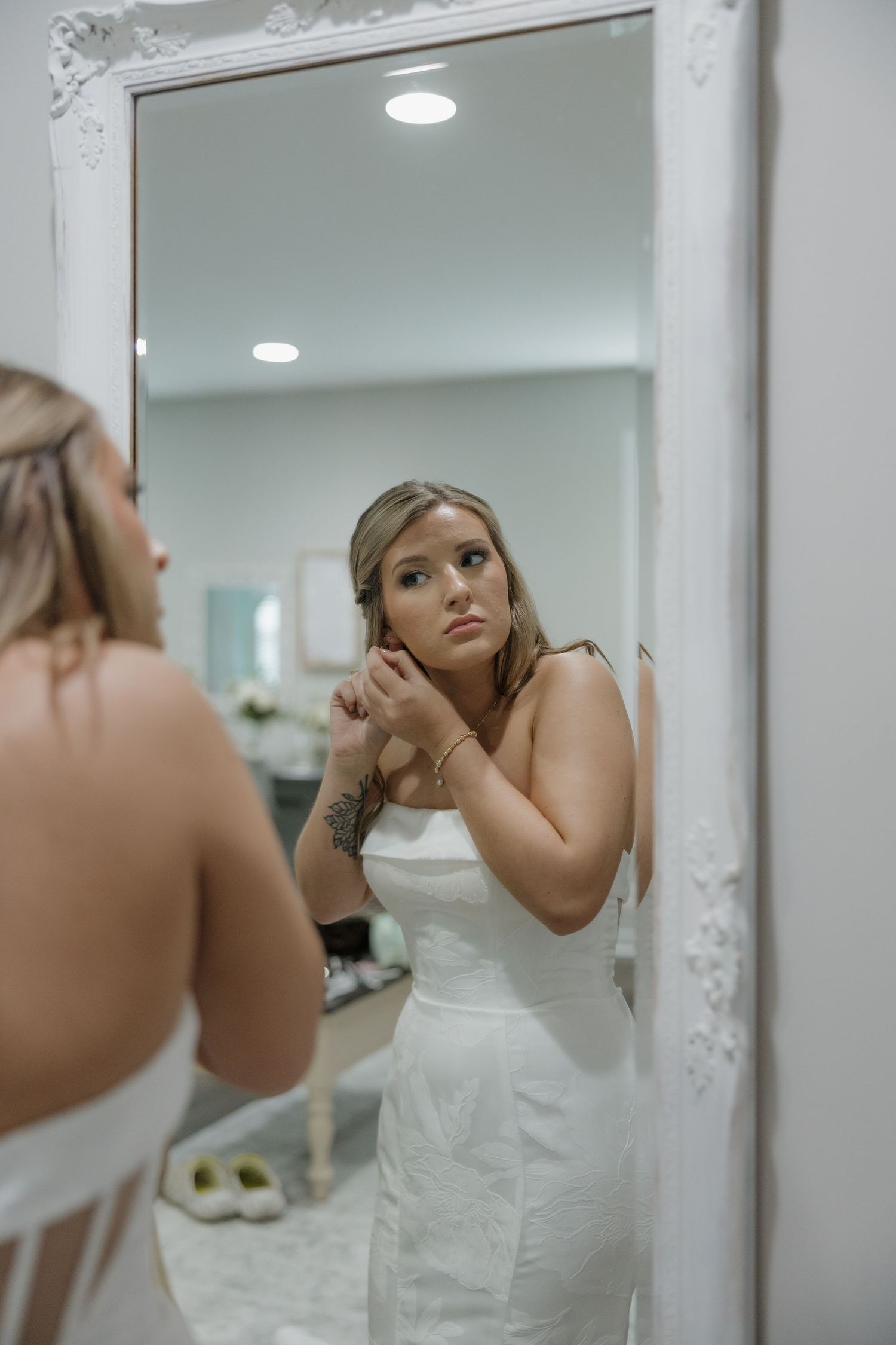 A woman in a white dress adjusts her earring while looking at her reflection in an ornate white-framed mirror.
