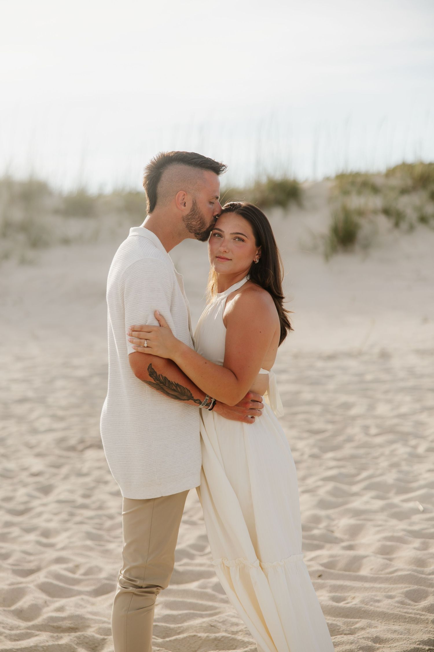 Couple embracing on sandy beach at golden hour, woman in white dress smiling at camera while man kisses her forehead.