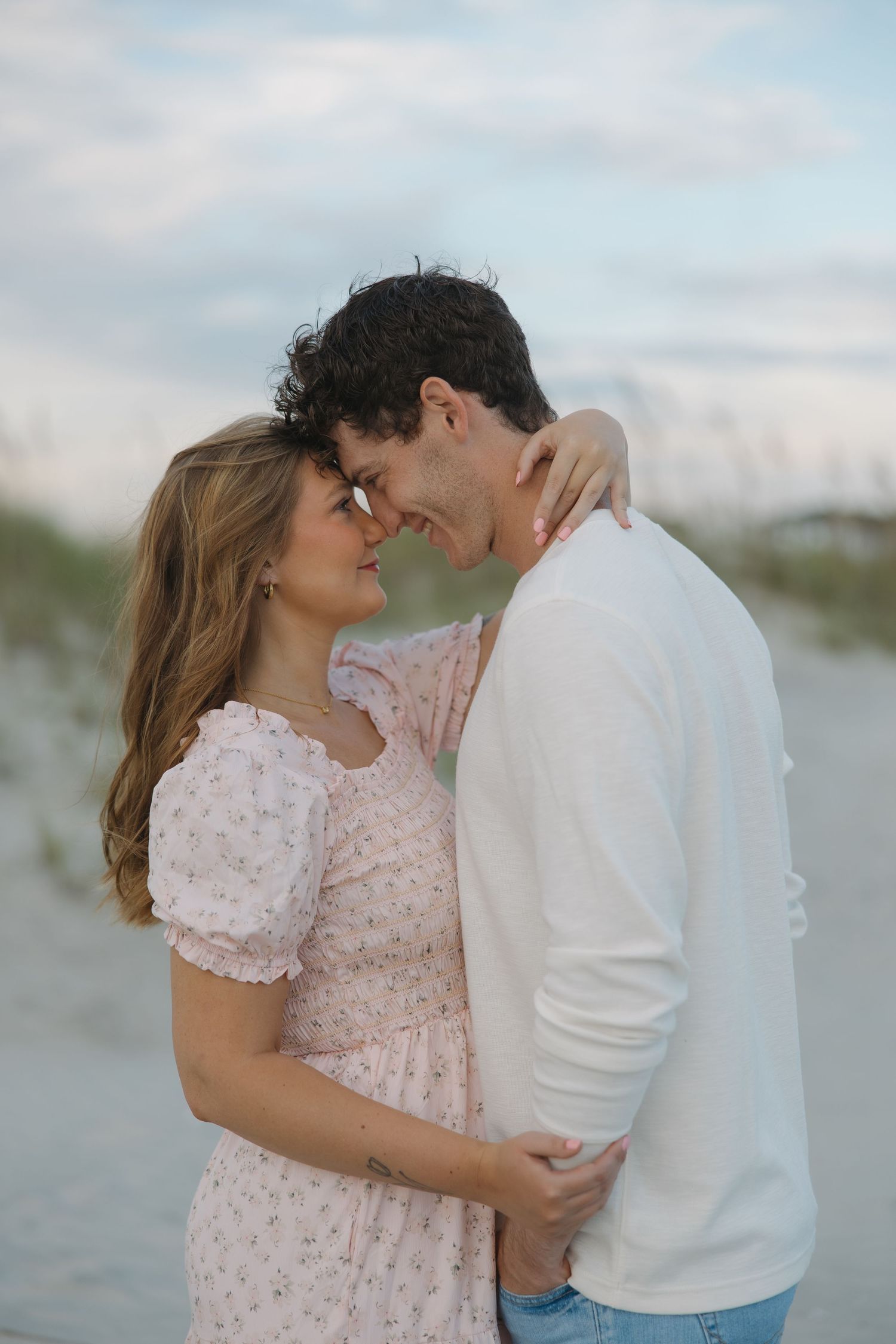 Romantic couple embracing on a beach at sunset, woman in floral dress and man in white sweater touching foreheads.