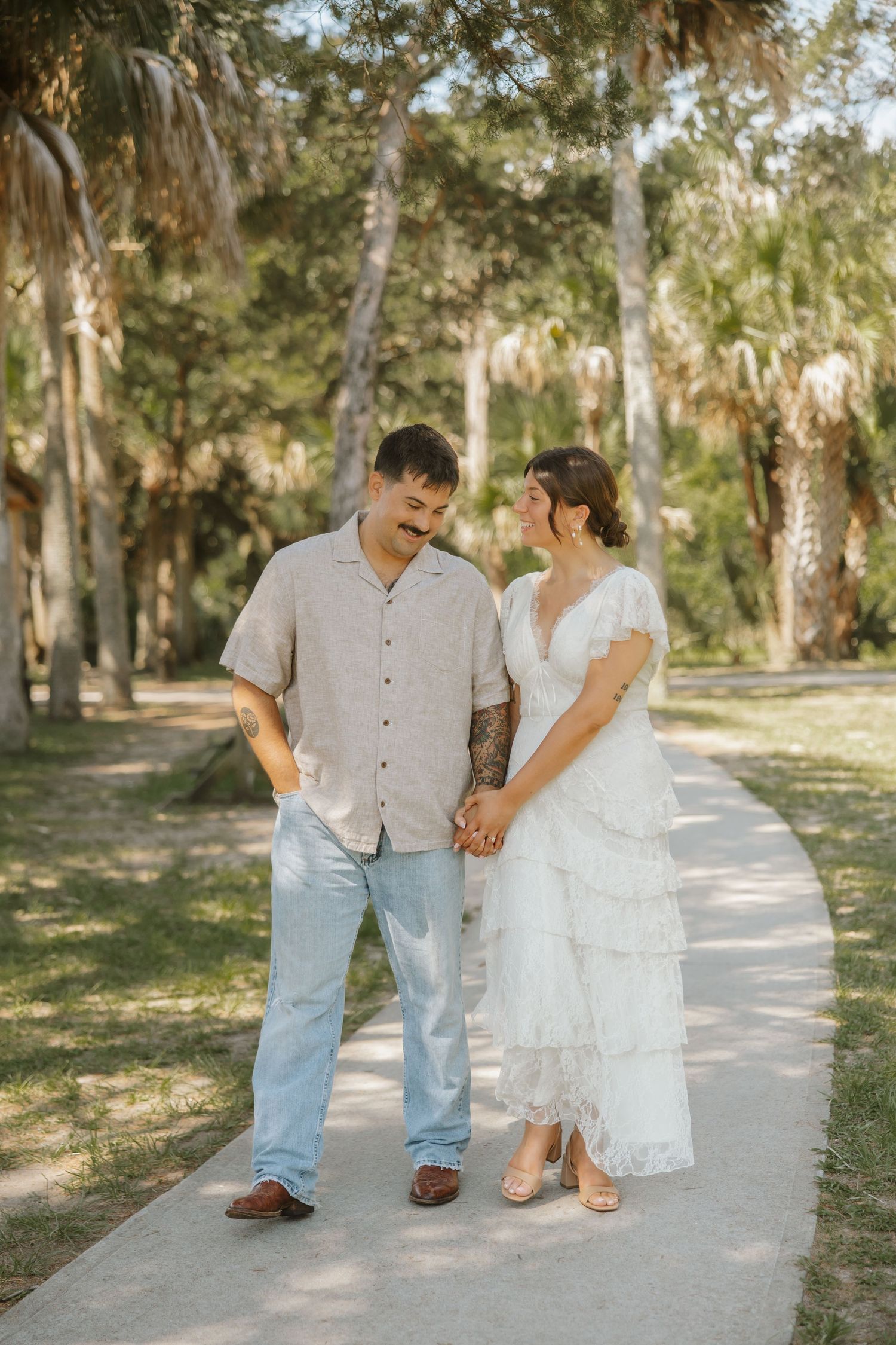 A couple in casual wedding attire holds hands while walking on a shaded park pathway surrounded by tall palm trees.