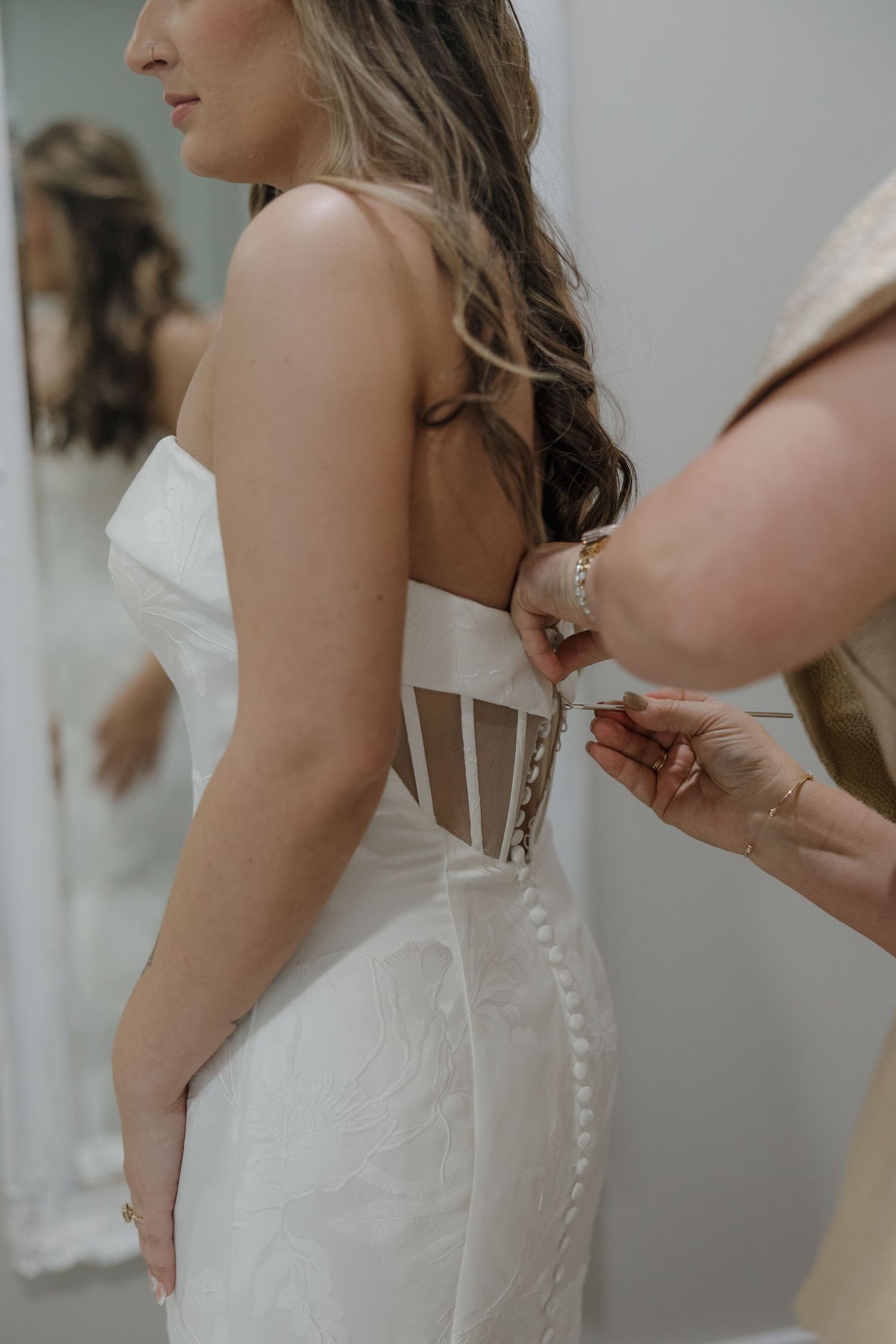 Bride in a strapless white lace-up corset wedding gown with a mirror reflection as a helper fastens the back buttons.