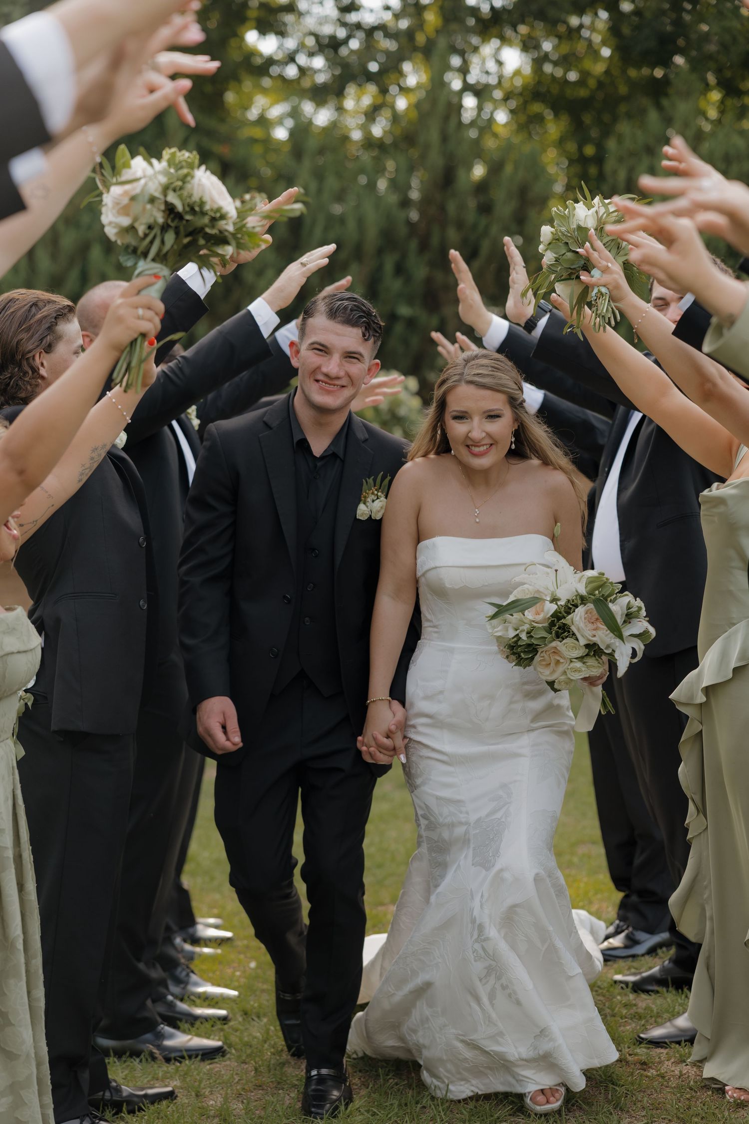 Smiling bride in a strapless white gown and groom in a black suit walk hand in hand through cheering wedding guests on a lawn.