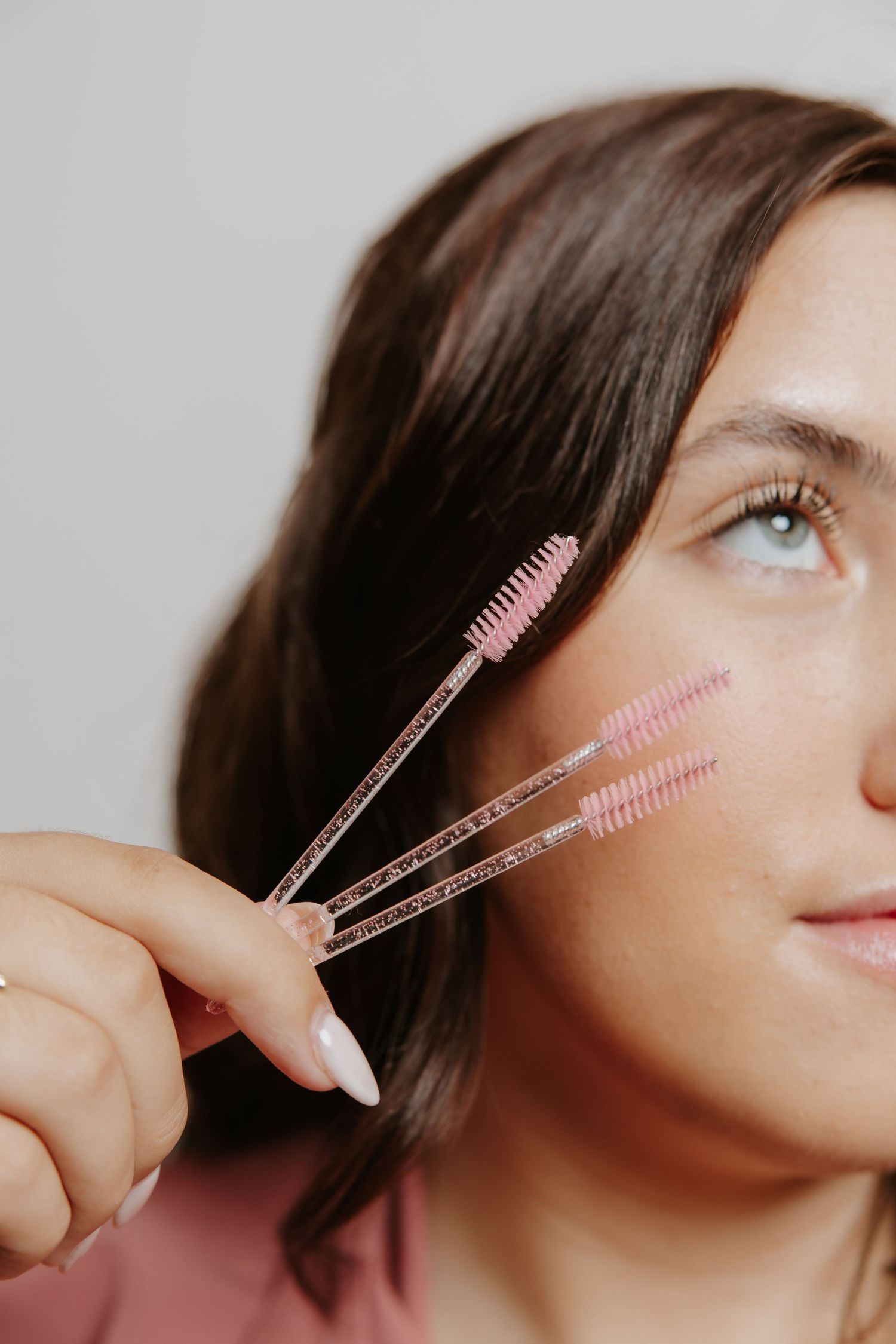 Woman holding three pink mascara wands near her face against a light gray background.