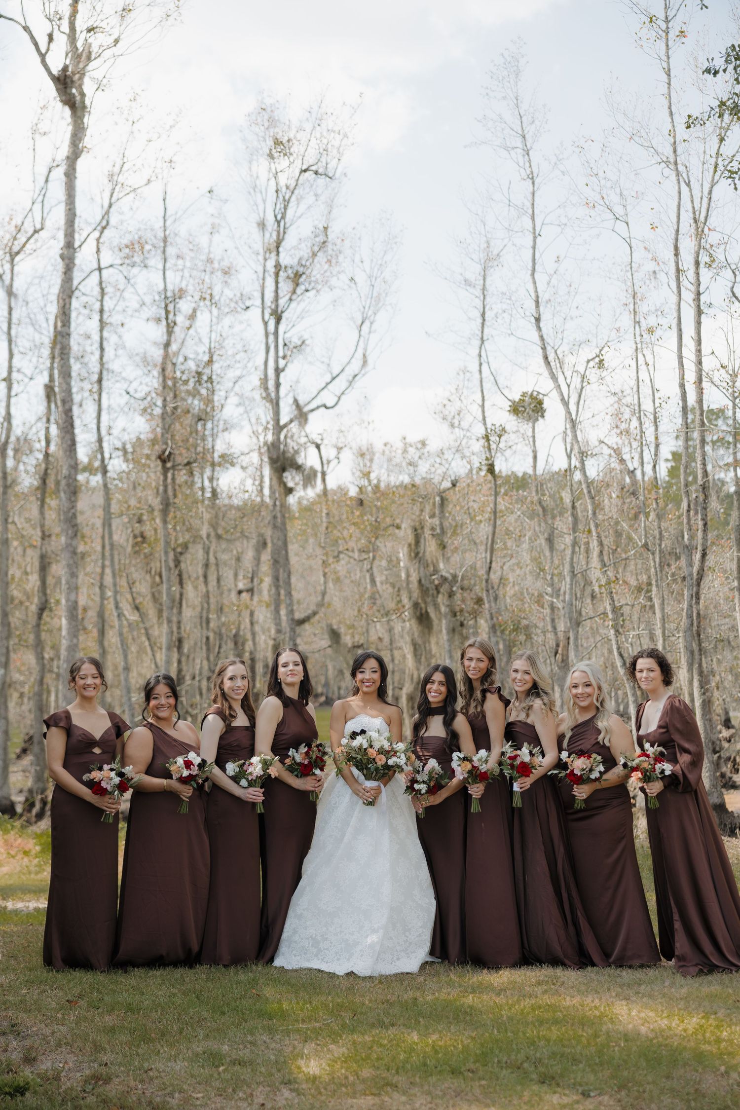 A bride in a white gown stands among bridesmaids in brown dresses holding bouquets in a wooded setting.