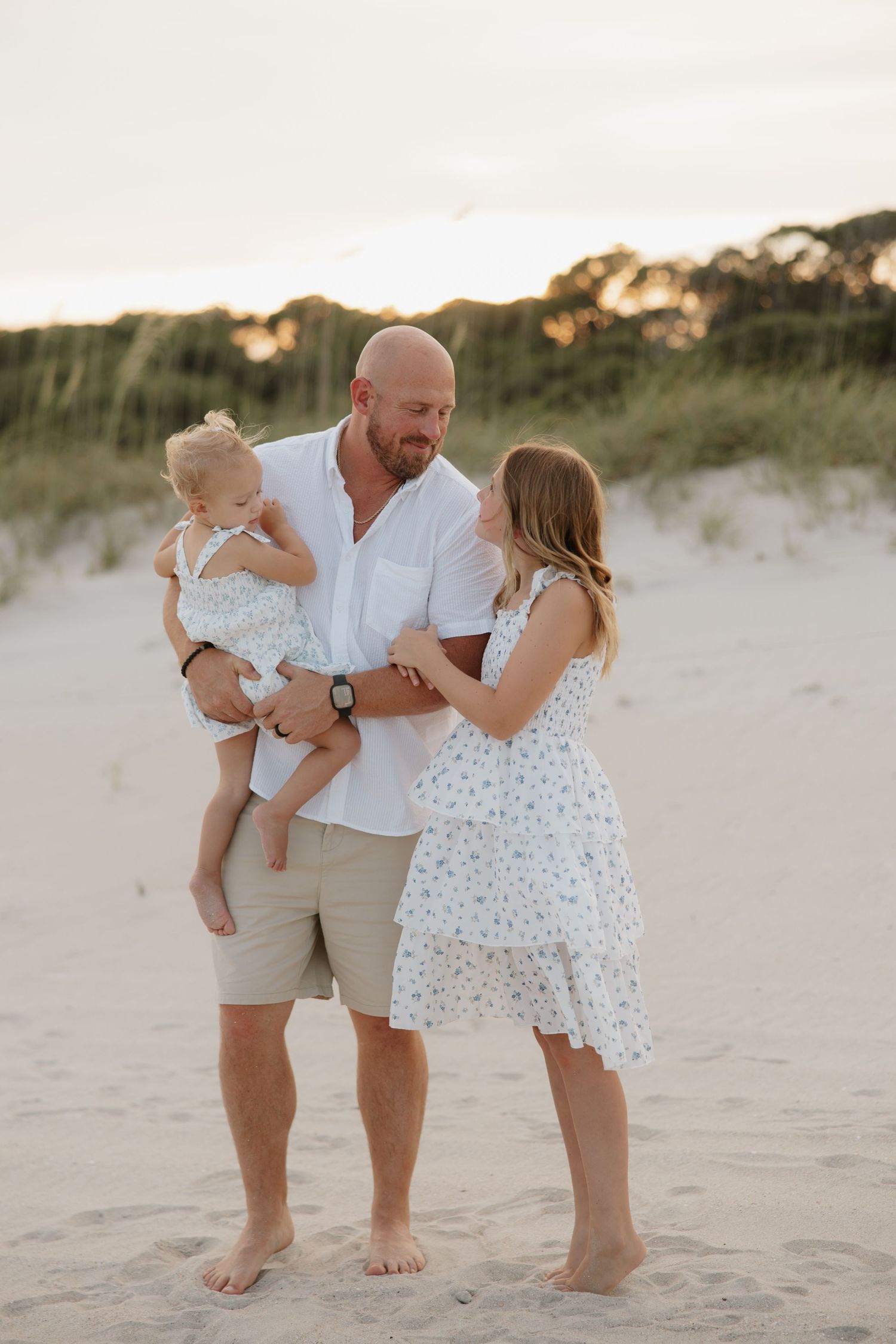 A family of three poses on a sandy beach at sunset, father holding baby while mother in floral dress smiles warmly.