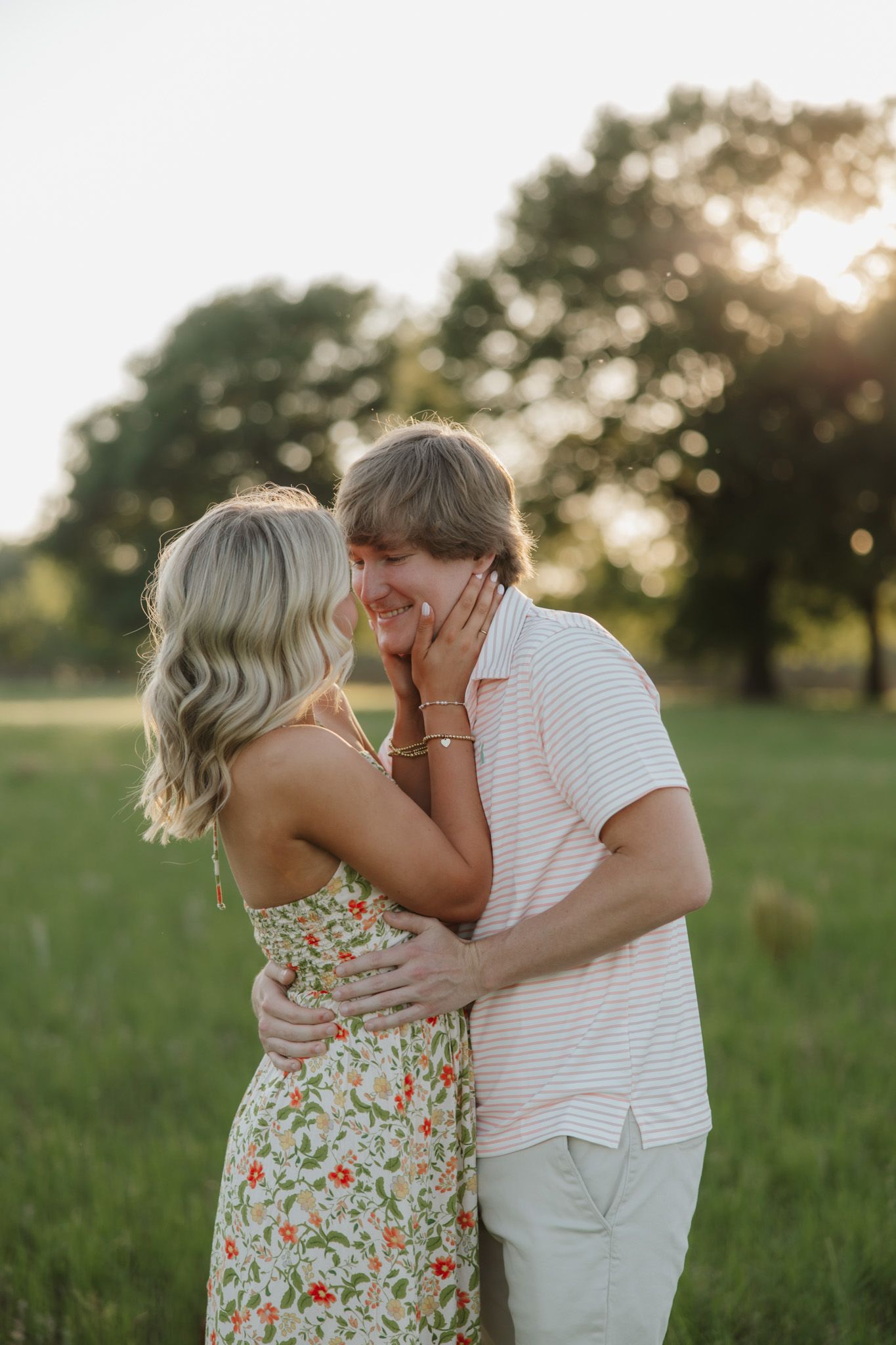 A couple shares a tender moment in a sunlit green field, the woman in a floral dress touching the mans smiling face.