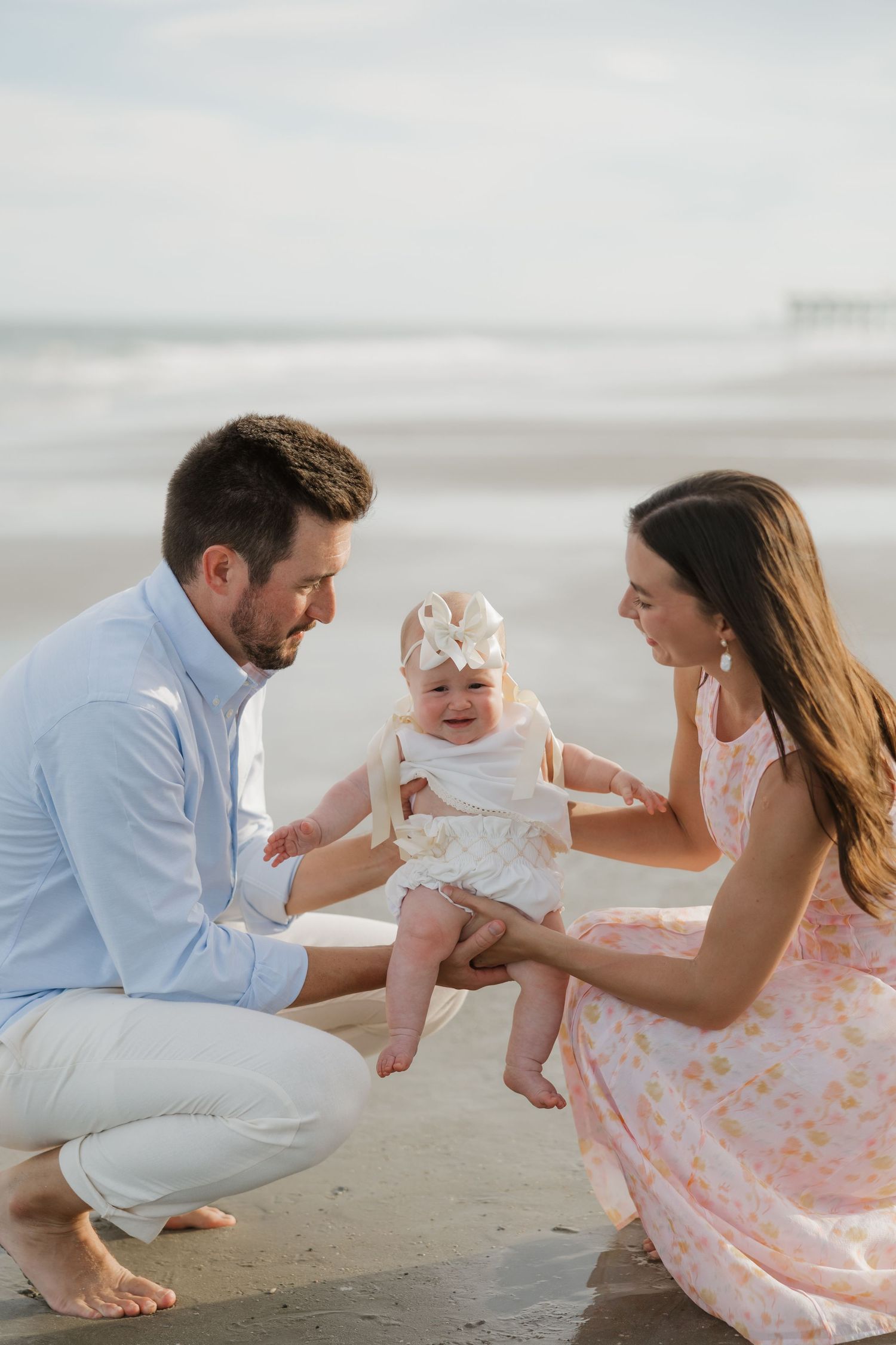 Young family with baby girl enjoys a sunny beach day, parents smiling at infant in white dress with bow headband.