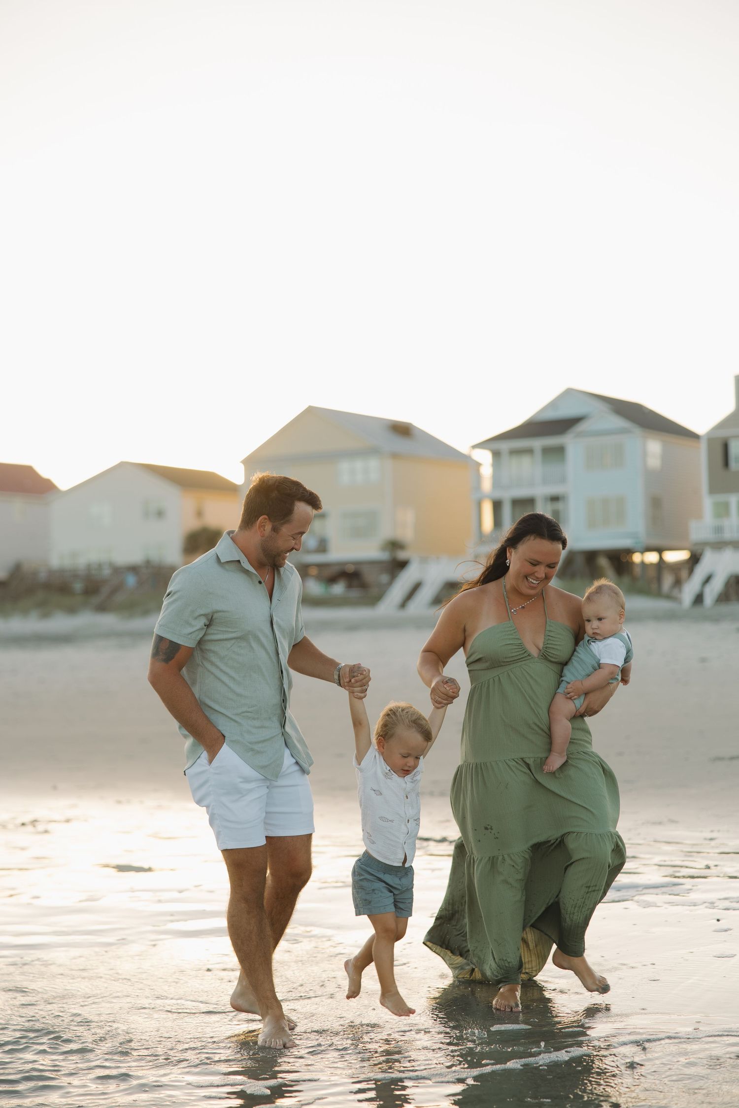 Family of four walking on beach at sunset, parents holding toddler's hands while mother carries baby in green dress.